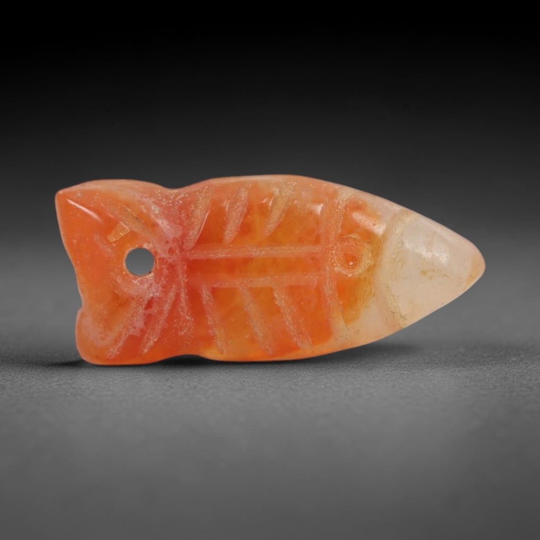 Ancient Carnelian Fish Amulet - Egyptian Figurine, 43AJ: Carved from translucent orange carnelian with a pale terminal tip, this amulet is formed as a stylized fish with incised linear detailing across the body to denote a central spine and radiating fin ra