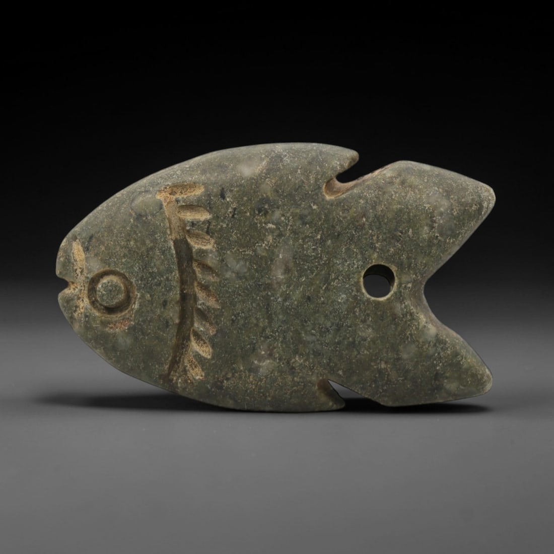Ancient Stone Fish - Egyptian Amulet Pendant, 49XR: Carved from mottled green stone, this amulet depicts a stylized fish with incised details representing the eye, mouth, and fin rays, alongside a drilled hole for suspension. The form and iconography a
