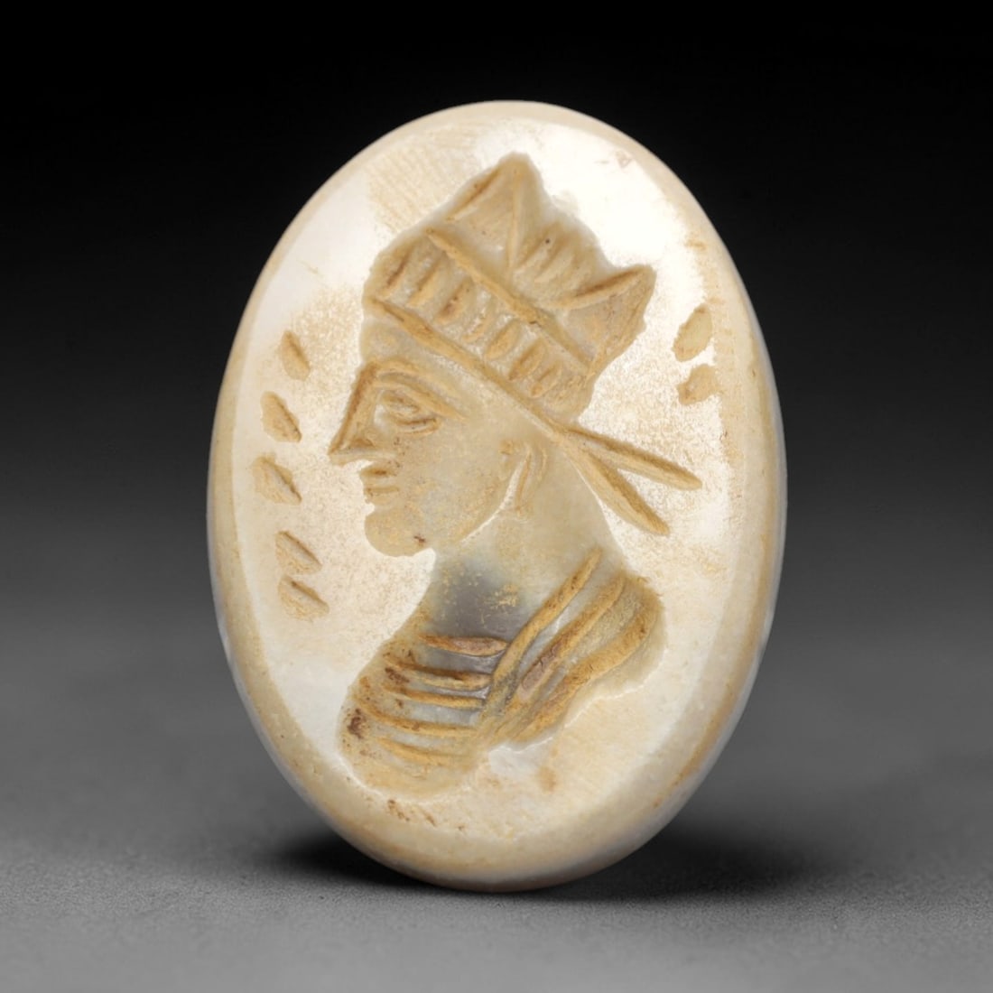 Ancient Agate Intaglio Bead - Near Eastern Figural Bust, 23ZW: Carved from pale banded agate, this oval intaglio bead features a recessed profile depiction of a figural bust adorned with a stylized headdress, flanked by incised decorative elements, characteristic