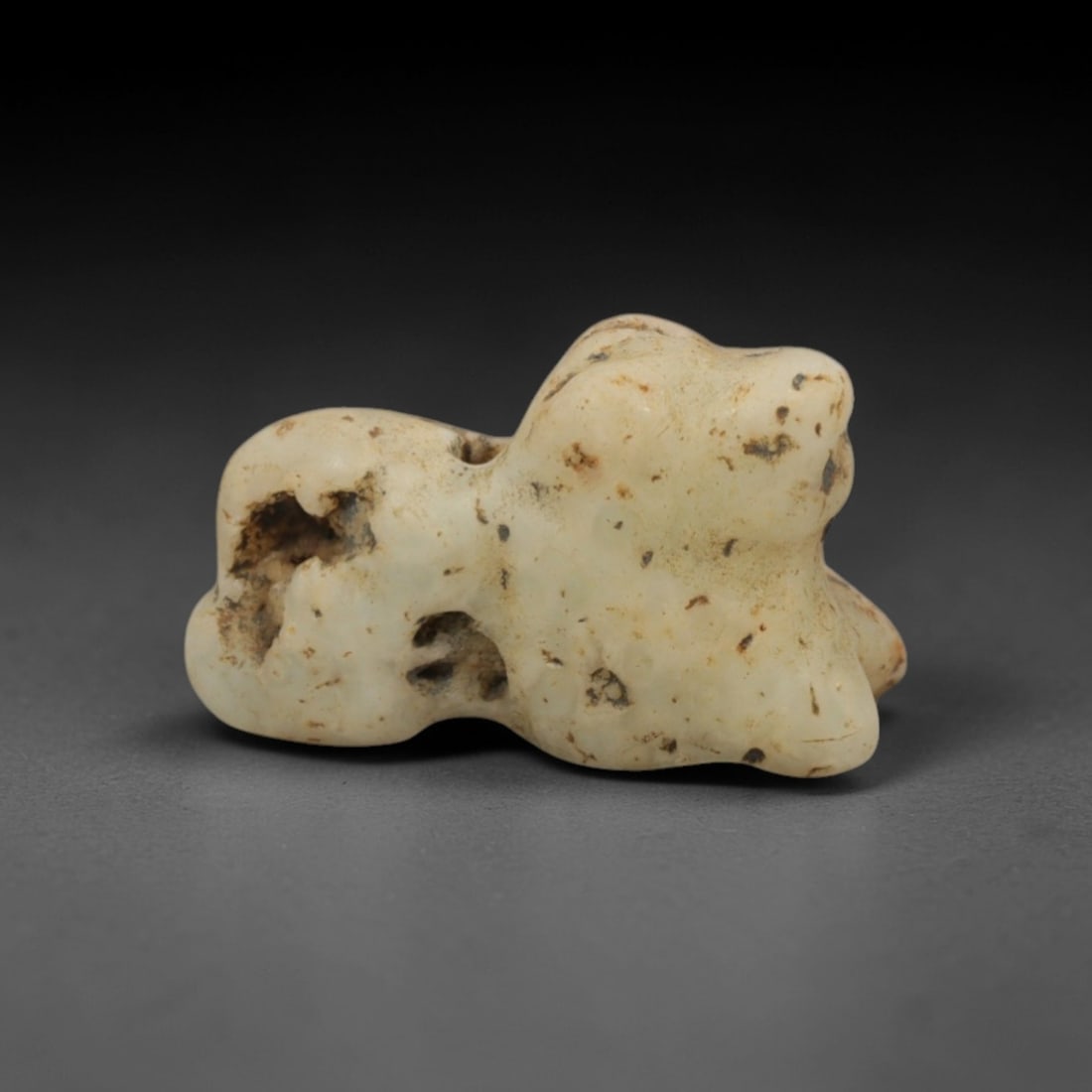 Ancient Cream Stone Zoomorphic Amulet - Near Eastern Miniature Figurine, 92XP: Carved from a weathered, creamy-white stone with natural grey and brown mineral inclusions, this miniature zoomorphic amulet depicts a stylized recumbent animal, likely a feline or small ungulate, a c