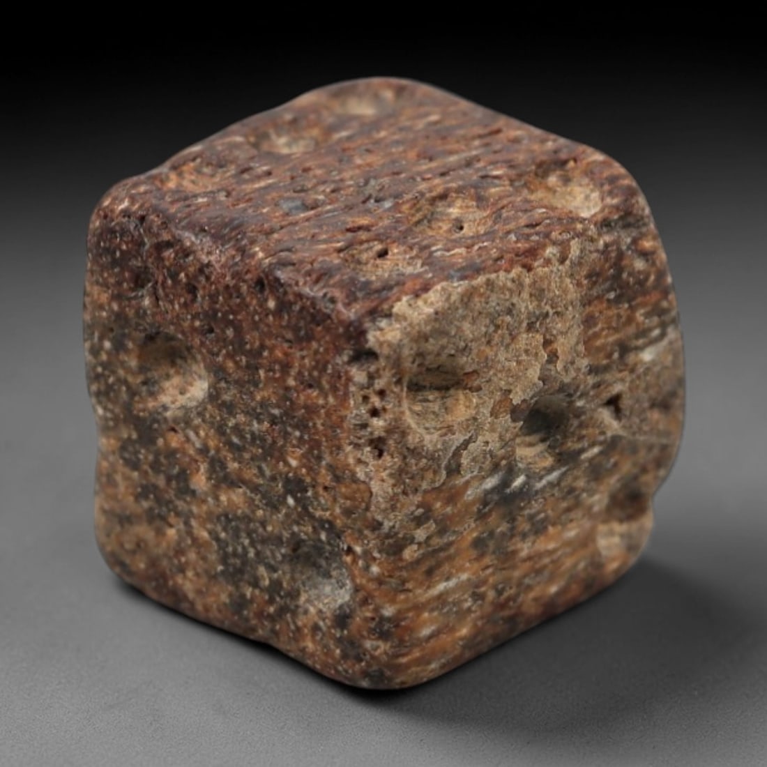 A Roman Stone Gaming Piece, Circa 1st-3rd Century CE, 9FT: Carved from granular stone with a predominantly reddish-brown matrix and lighter mineral inclusions, the object is shaped as a roughly hewn cube with rounded edges and slightly irregular faces. The su