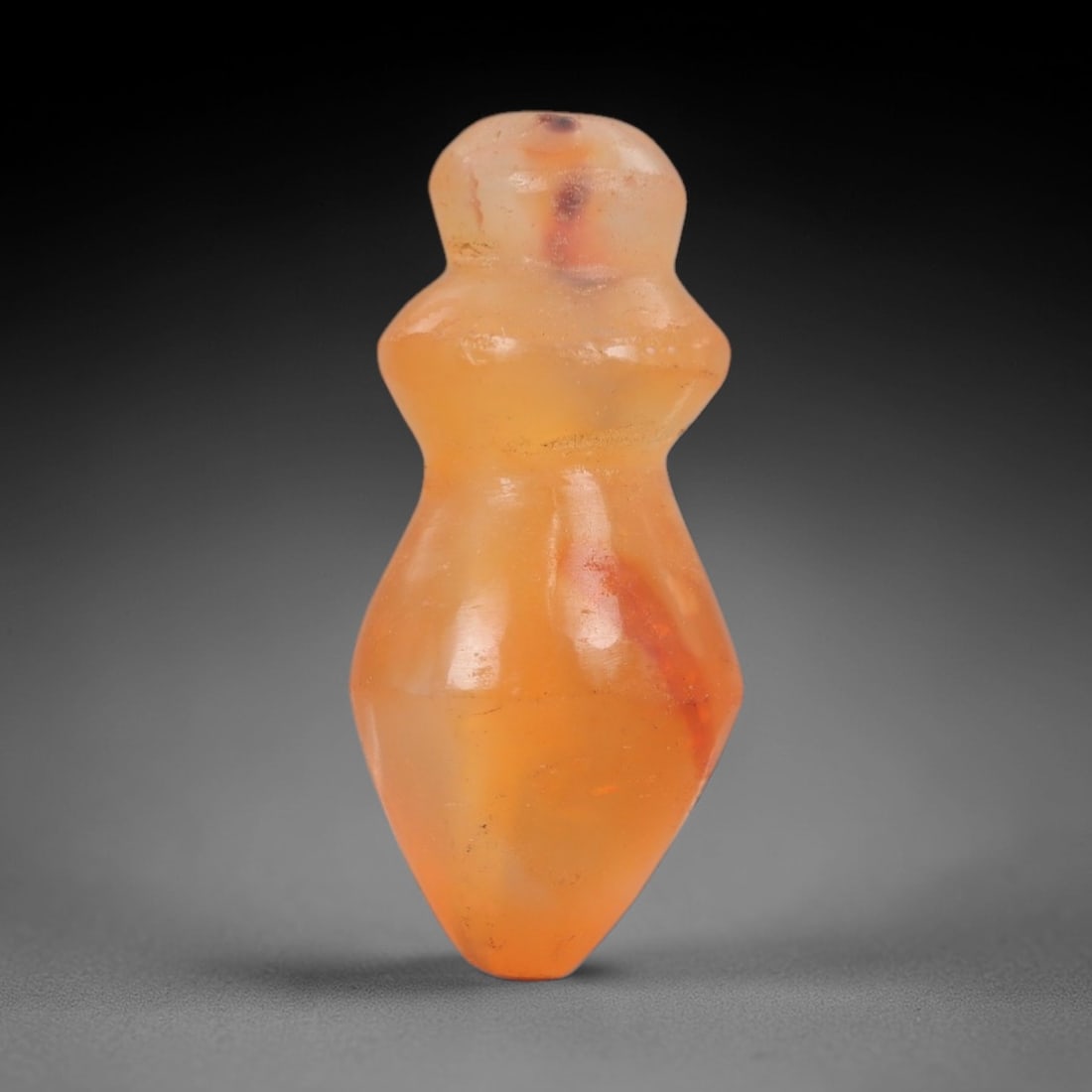 Ancient Carnelian Fertility Amulet - Near Eastern Figurine, 59AJ: Carved from translucent orange carnelian, this amulet is formed as a stylized anthropomorphic fertility figure with a rounded head, narrow neck, and bulbous lower body tapering to a pointed base, cons