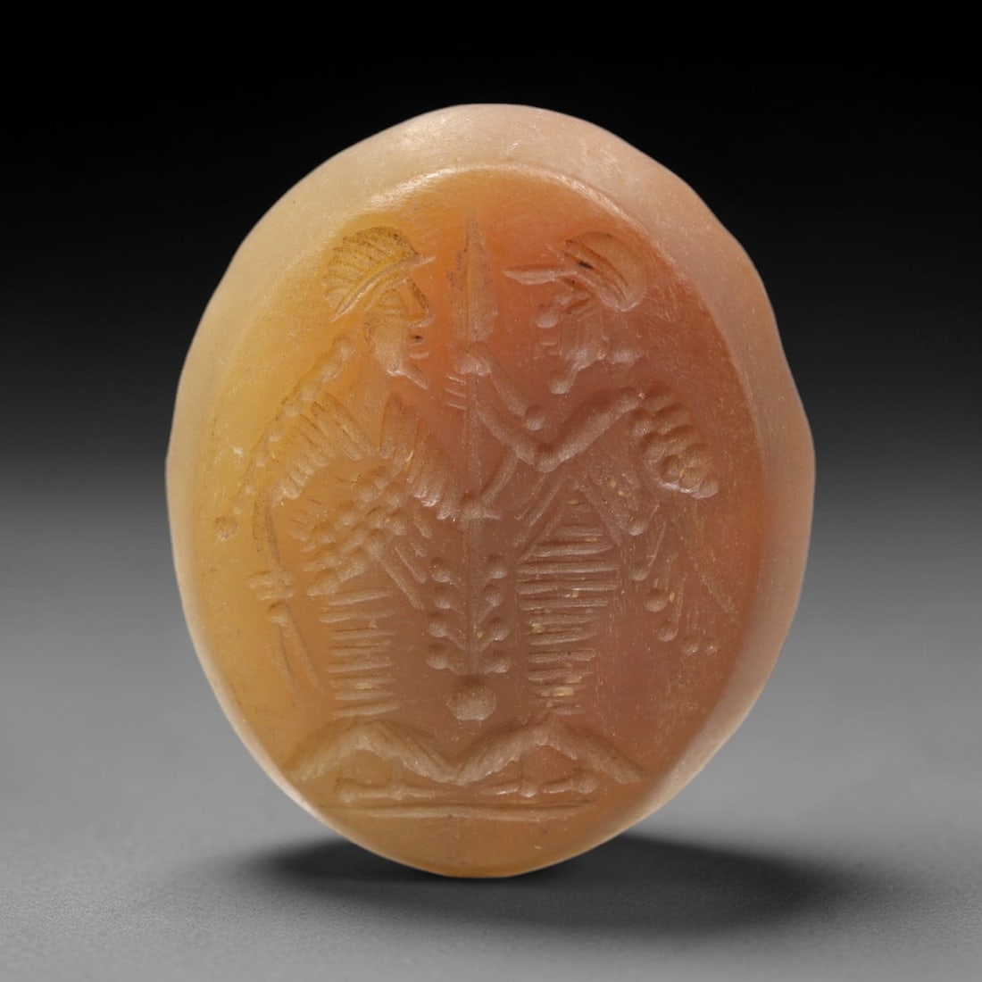 Ancient Carnelian Intaglio - Dual Warrior Figure Seal, 38XV: Crafted from polished warm-hued carnelian, this substantial oval intaglio seal depicts two confronting warrior figures in profile, each adorned with crested helmets, layered torsos, and holding uprigh