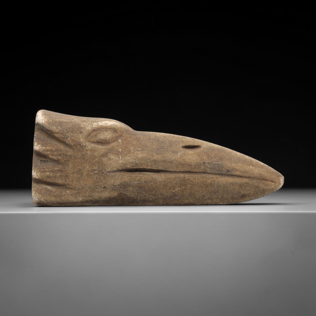 Ancient Stone Zoomorphic Bird Head - Near Eastern Amulet, 79ZV: This artifact is a carved stone zoomorphic bird head amulet, crafted from a granular, earth-toned stone consistent with ancient Near Eastern lithic traditions. The relief carving depicts a stylized av