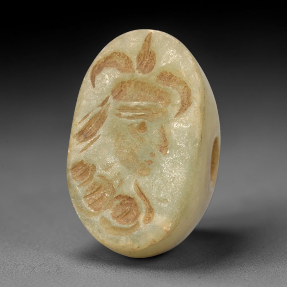 Ancient Serpentine Stamp Seal - Near Eastern Anthropomorphic Bust, 65ZR: Carved from pale green serpentine, this hemispherical stamp seal features a raised relief depiction of a profile anthropomorphic bust, adorned with a horned headdress and stylized facial details, alon