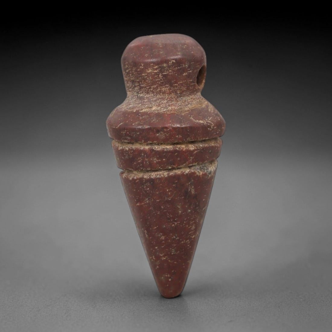 Ancient Stone Cone Amulet - Near Eastern Carved Pendant, 43AG: This amulet is carved from a rich reddish stone with a granular texture and weathered, earthy patina. The form is a tapered cone, with a rounded upper segment featuring a lateral drilled suspension ho