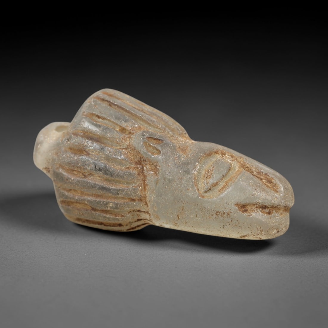 Ancient Glass Feline Head - Zoomorphic Amulet Figurine, 18AL: This zoomorphic amulet is carved from translucent pale glass, shaped into the stylized head of a feline creature with incised linear details defining the mane, eye, and muzzle. A suspension hole at th