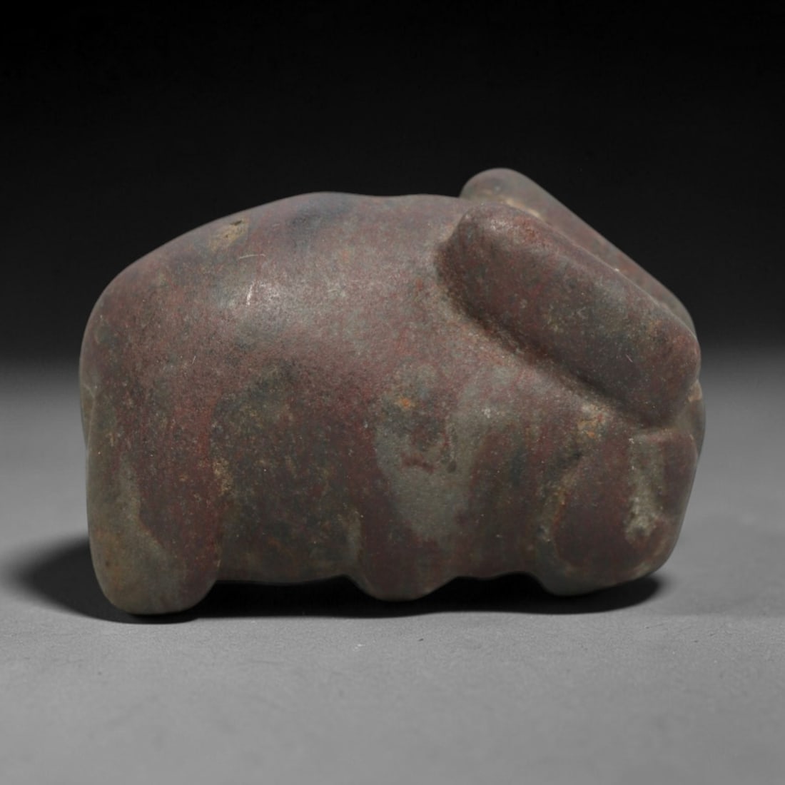 Ancient Stone Hare Amulet - Near Eastern Animal Figure, 38ZV: This amulet is carved from a mottled dark red and grey stone, depicting a compact recumbent hare with stylized ears and subtle incised details defining the facial features. A small perforation is dril