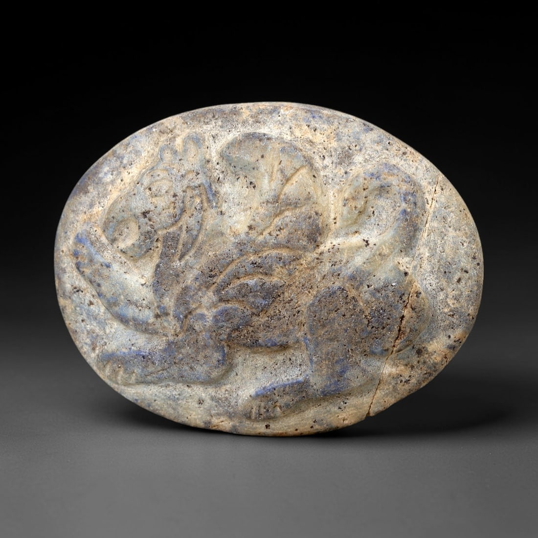 Ancient Lapis Animal Plaque - Near Eastern Relief Amulet, 67AM: This oval plaque is carved from lapis lazuli, displaying a mottled blue-grey patina with natural mineral inclusions consistent with ancient Near Eastern lapis working. The raised relief depicts a styl