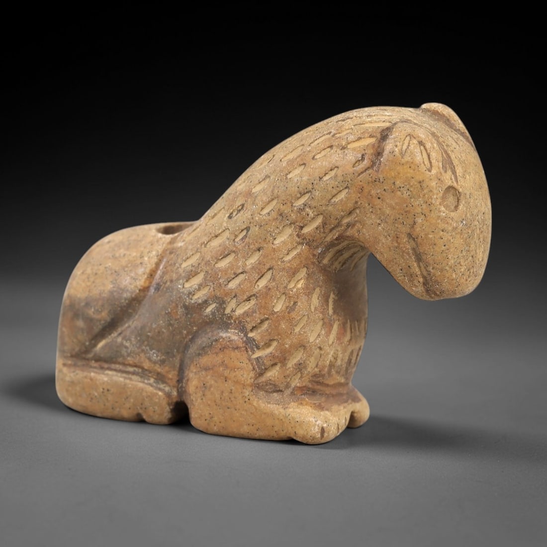 Ancient Stone Animal Amulet - Near Eastern Figurine, 86AL: Carved from tan speckled stone, this amulet depicts a recumbent quadrupedal animal, likely a canine or feline, with incised linear patterns across its body to denote fur. The head is rounded with a si