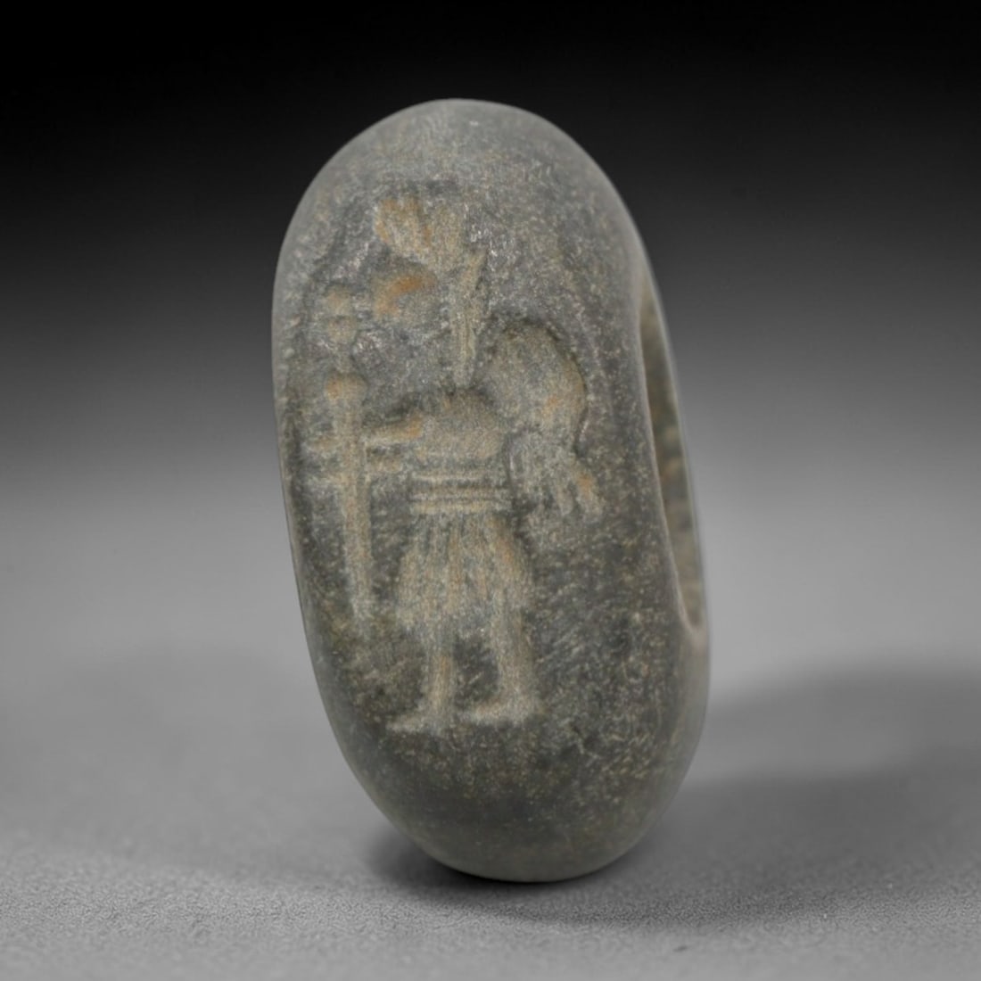 Ancient Stone Hemispherical Stamp Seal - Near Eastern Warrior Figure, 4ZT: Crafted from dense, fine-grained dark stone, this hemispherical stamp seal bears precise incised carving of a standing warrior figure, adorned with a headdress, holding a staff or weapon, and clad in