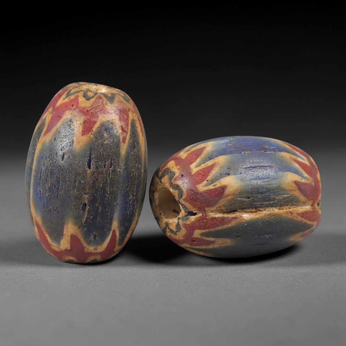 A Pair of Venetian Glass Chevron Beads, 18th-19th Century, 75AC: Crafted from translucent glass with a pale yellow core, each bead is decorated with a chevron pattern of alternating deep blue and vibrant red layers, radiating from the central perforation to the out