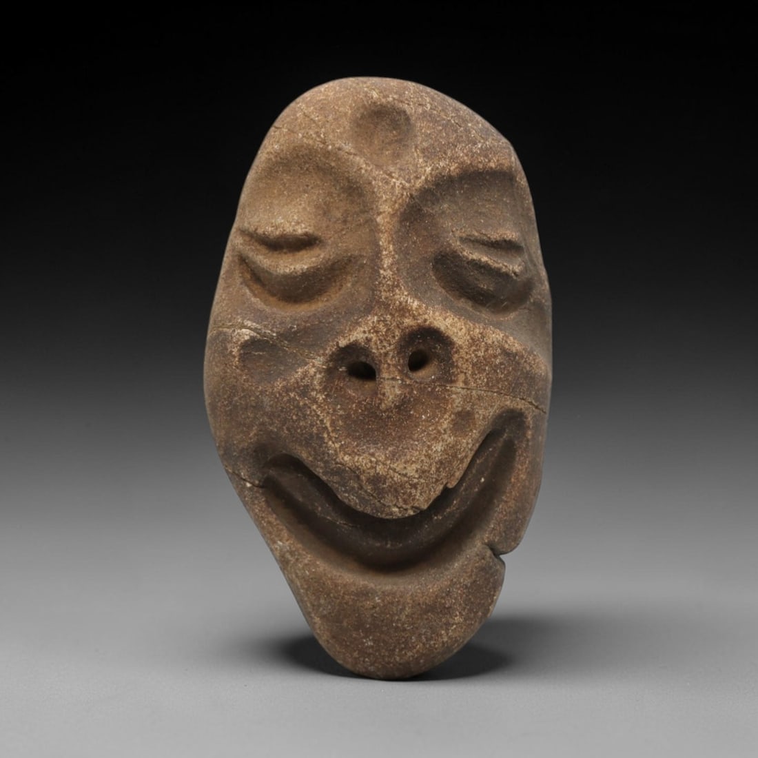 Ancient Stone Anthropomorphic Face - Near Eastern Amulet, 74ZV: This artifact is a carved stone anthropomorphic face amulet, rendered in a granular, earth-toned stone consistent with ancient Near Eastern lithic traditions. The relief carving depicts a stylized fac