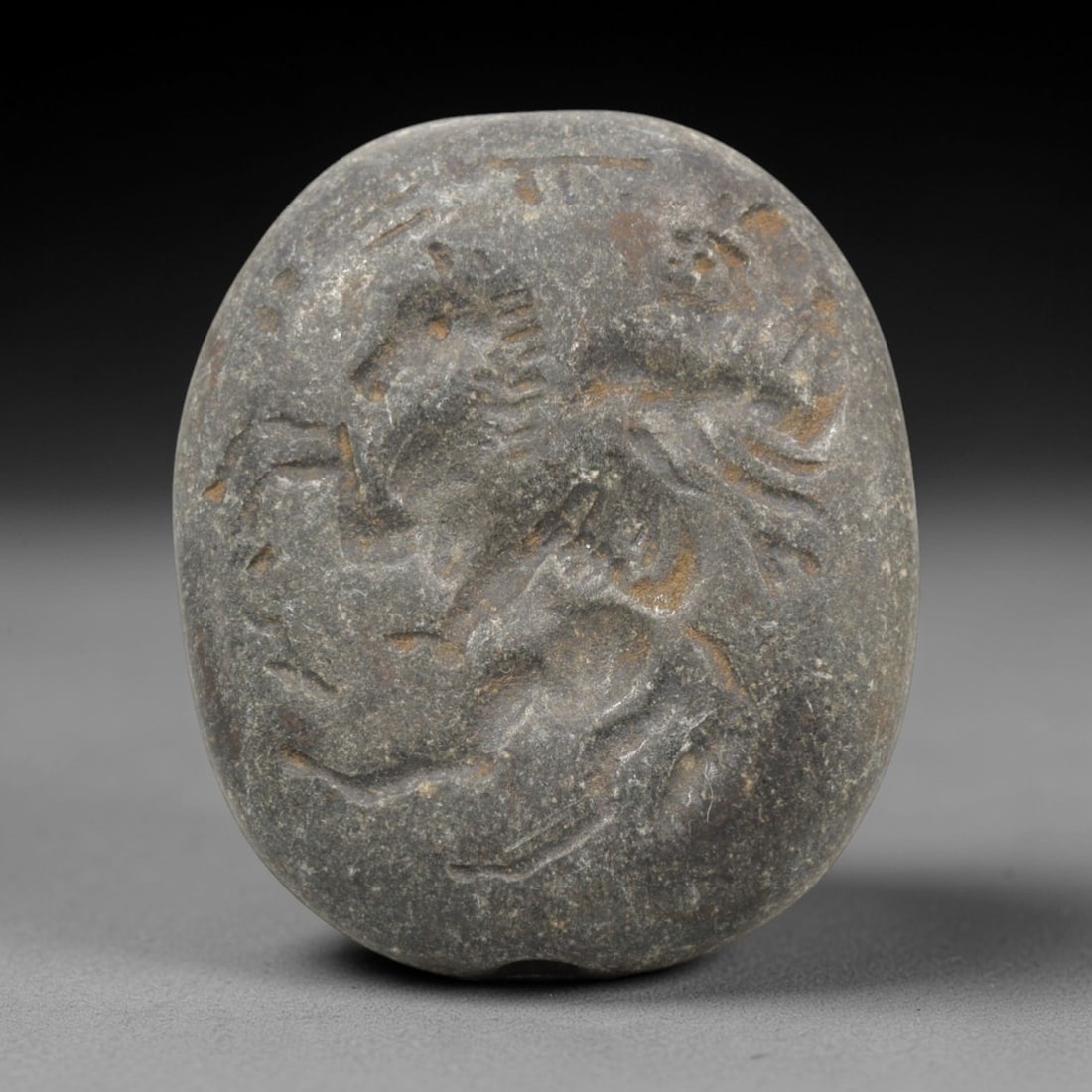 Ancient Stone Intaglio Bead - Near Eastern Warrior on Horse, 76ZR: Carved from a dense, dark stone, this oval intaglio bead depicts a warrior mounted on horseback in high relief. The composition captures the dynamic posture of the horse in motion, with the warrior's