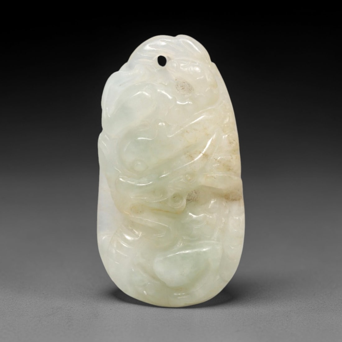 A Chinese Jadeite Jade Dragon Pendant, 20th Century, 61FR: Carved from pale translucent jadeite jade with natural faint brownish tonal variations, the pendant has an oval form with a drilled suspension hole at the apex. It depicts a dragon rendered in high re