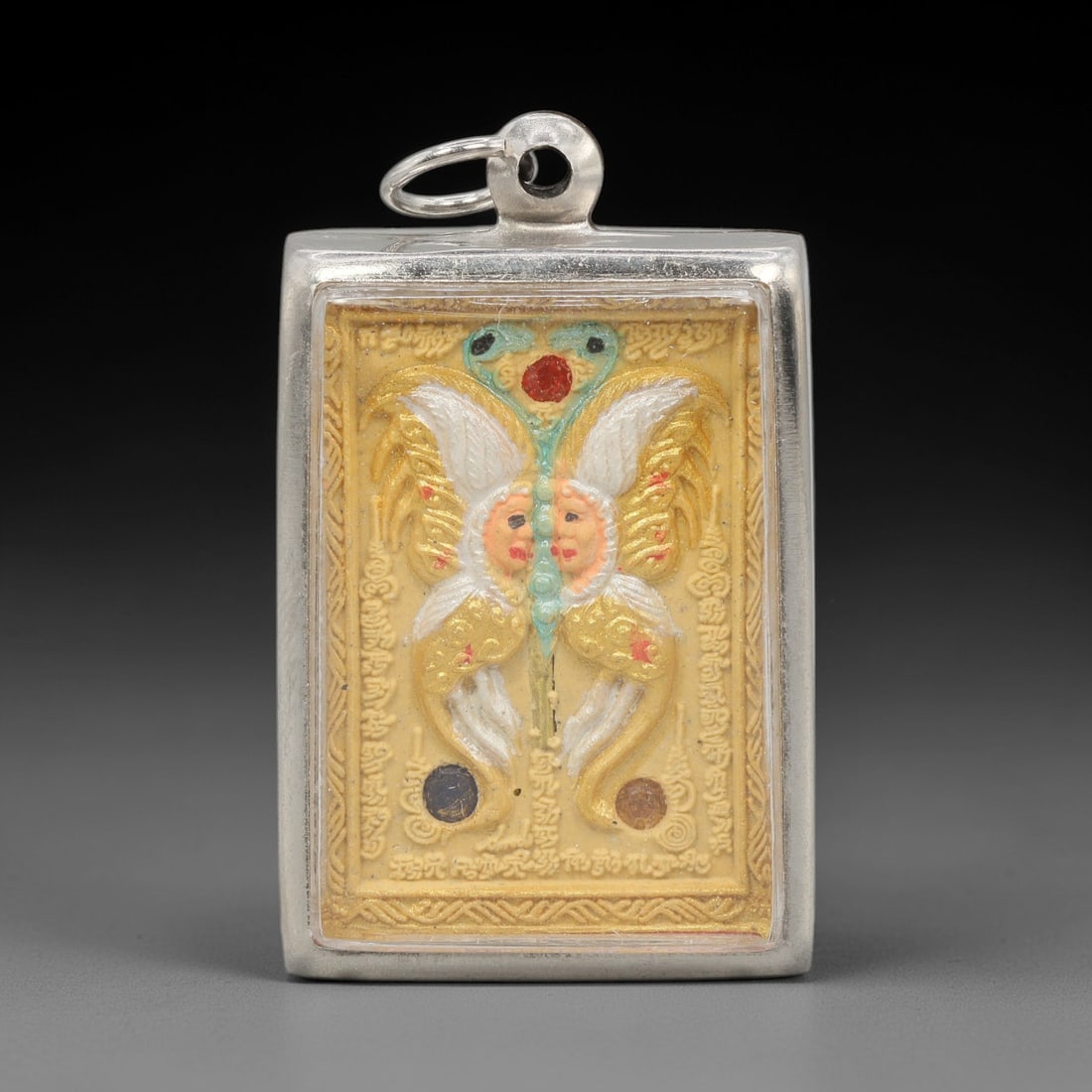 A Thai Butterfly Amulet in Silver Frame, Late 20th Century, 65ZH: A rectangular amulet, measuring 5 x 3 cm, set within a silver frame with a suspension loop. The obverse depicts a butterfly deity with two human faces, rendered in gold, white, and multicolored relief
