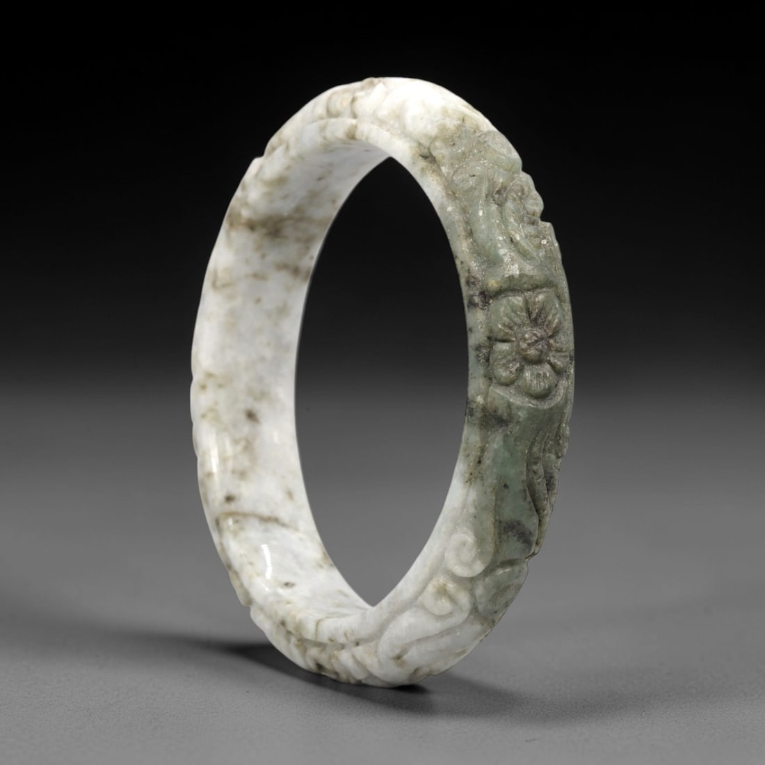 A Chinese Jadeite Jade Bangle Bracelet, 20th Century, 51FS: Carved from variegated jadeite jade with natural green, white, and greyish tonal variation, the bangle is formed as a continuous circular band. The exterior surface is decorated with raised relief car