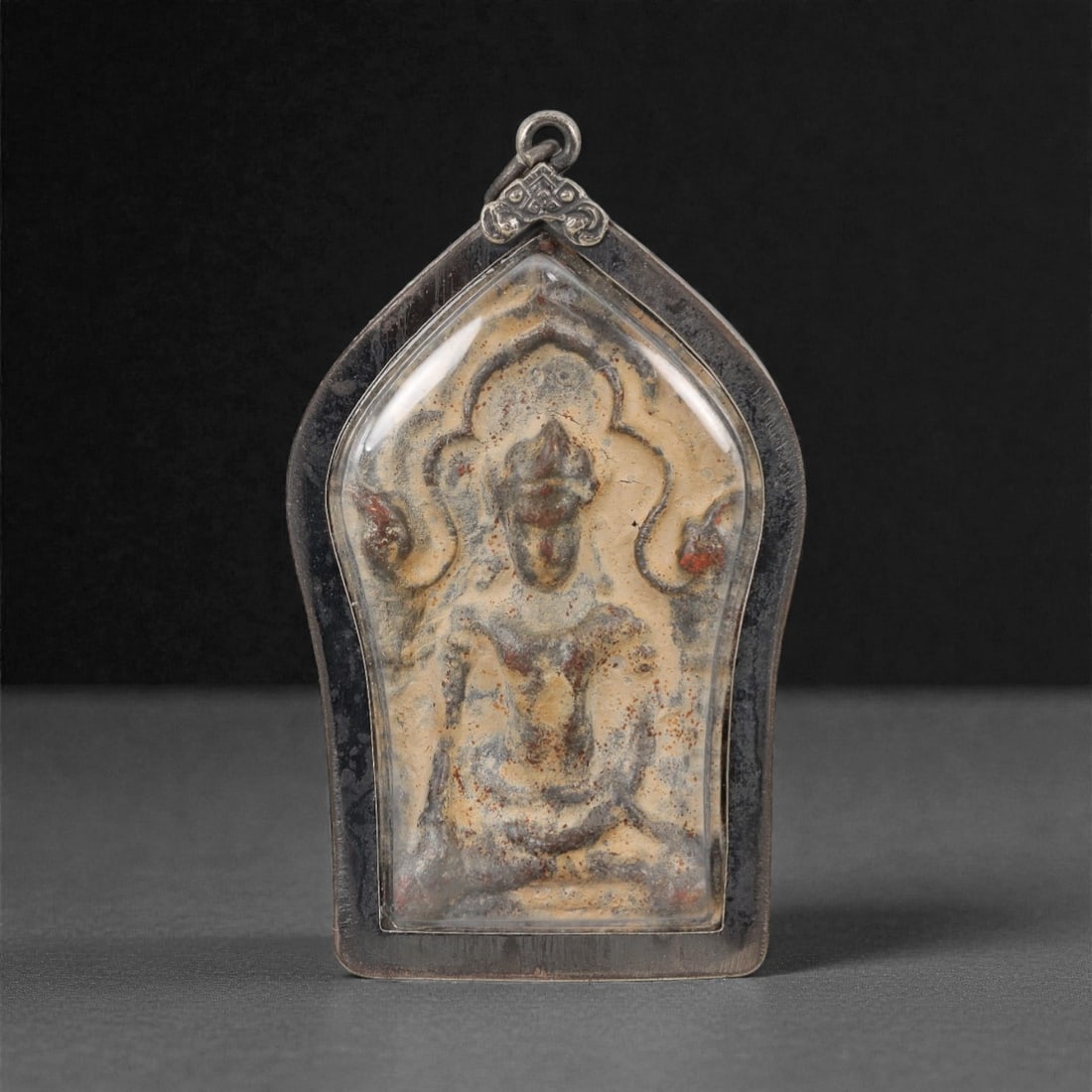 A Thai Seated Deity Amulet with Metal Frame, 20th Century, 31FP: The amulet is of shield-shaped form, featuring a molded seated deity figure on the obverse, rendered in a dark patinated material against a light beige ground. The deity is depicted with a multi-tiere