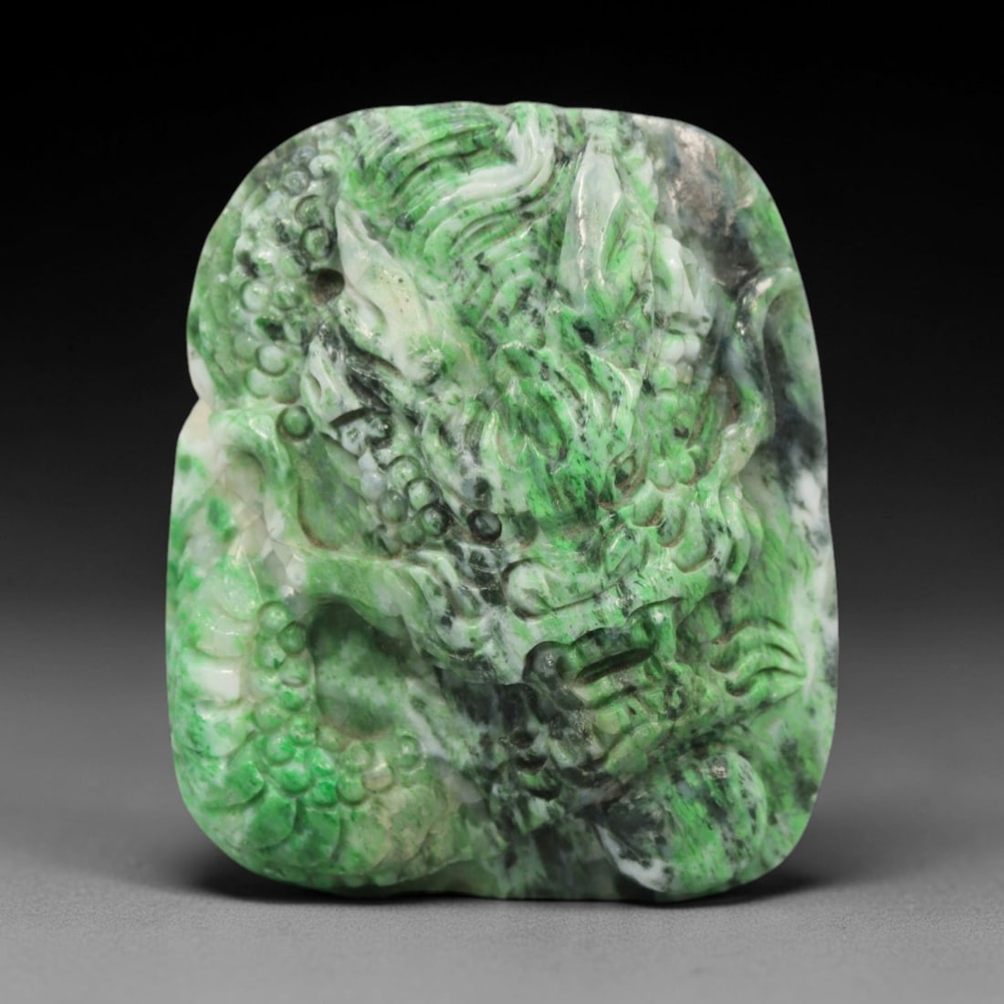 A Chinese Jadeite Jade Pendant, 20th Century, 14ZL: Carved from green jadeite jade with natural dark green and greyish inclusions distributed throughout the matrix, the pendant is formed as an irregularly rectangular object with a relief-carved dragon