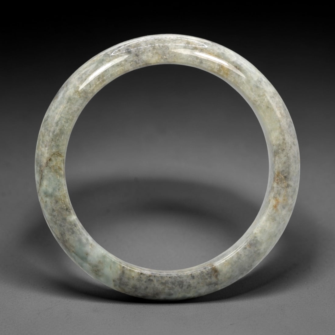 A Chinese Jadeite Jade Bangle Bracelet, 20th Century, 6ZL: Carved from jadeite jade with a translucent pale greenish-grey base and scattered russet-brown inclusions, the bangle is formed as a continuous circular band with a rounded cross-section. The surface