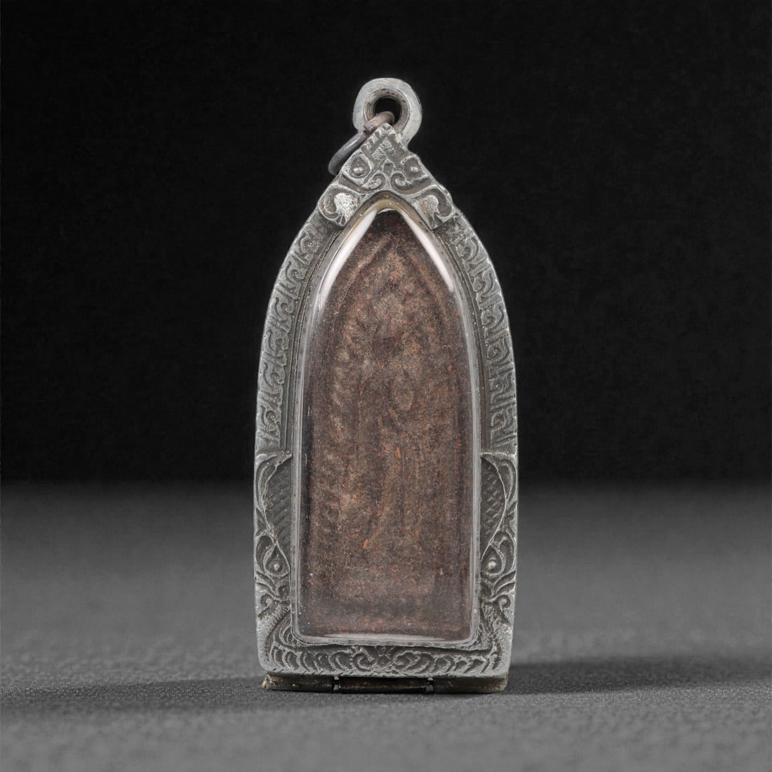 A Thai Clay Buddha Amulet in Metal Case, 20th Century, 36ZH: The amulet is formed from clay, with one side depicting a standing Buddha figure, and the reverse displaying a dark, textured surface with a linear marking. The amulet is enclosed within a pointed-arc