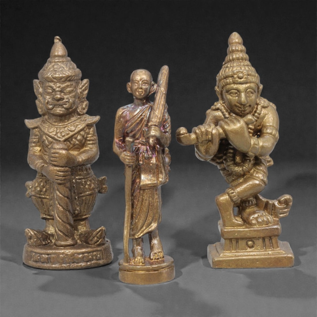 Three Thai Bronze Guardian and Monk Miniature Figures, 20th Century, 26ZH: Cast from bronze with a warm, aged patina, the set comprises three miniature figures: a fierce guardian figure (yaksha) with ornate armor and headdress, a standing monk holding an umbrella and staff,