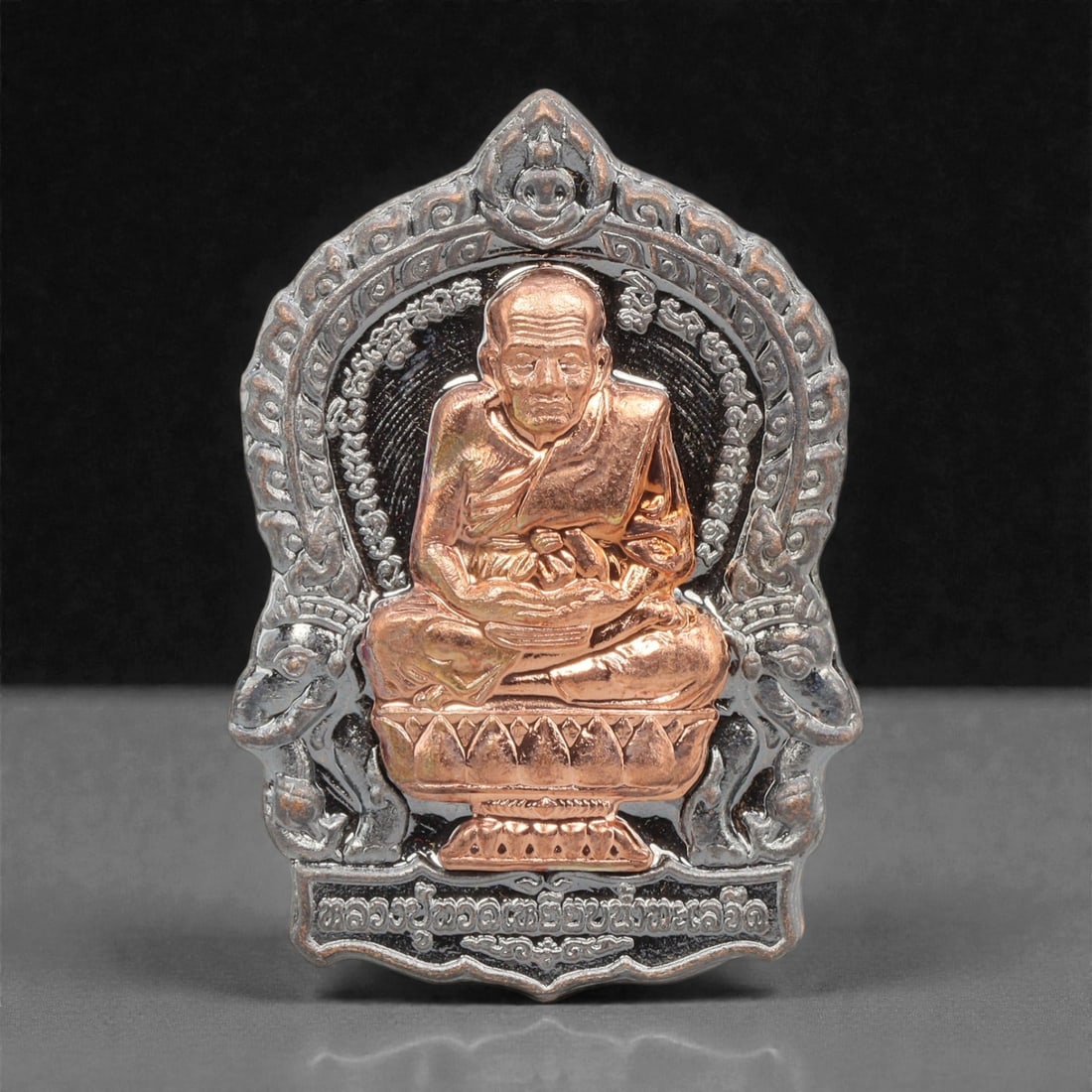 A Thai Bi-Metallic Monk Portrait Amulet, 21st Century, 82FP: Cast with a bi-metallic construction, the amulet has a shield-shaped form with a pointed upper apex. The obverse features a high-relief copper-toned bust portrait of a seated Buddhist monk, set agains