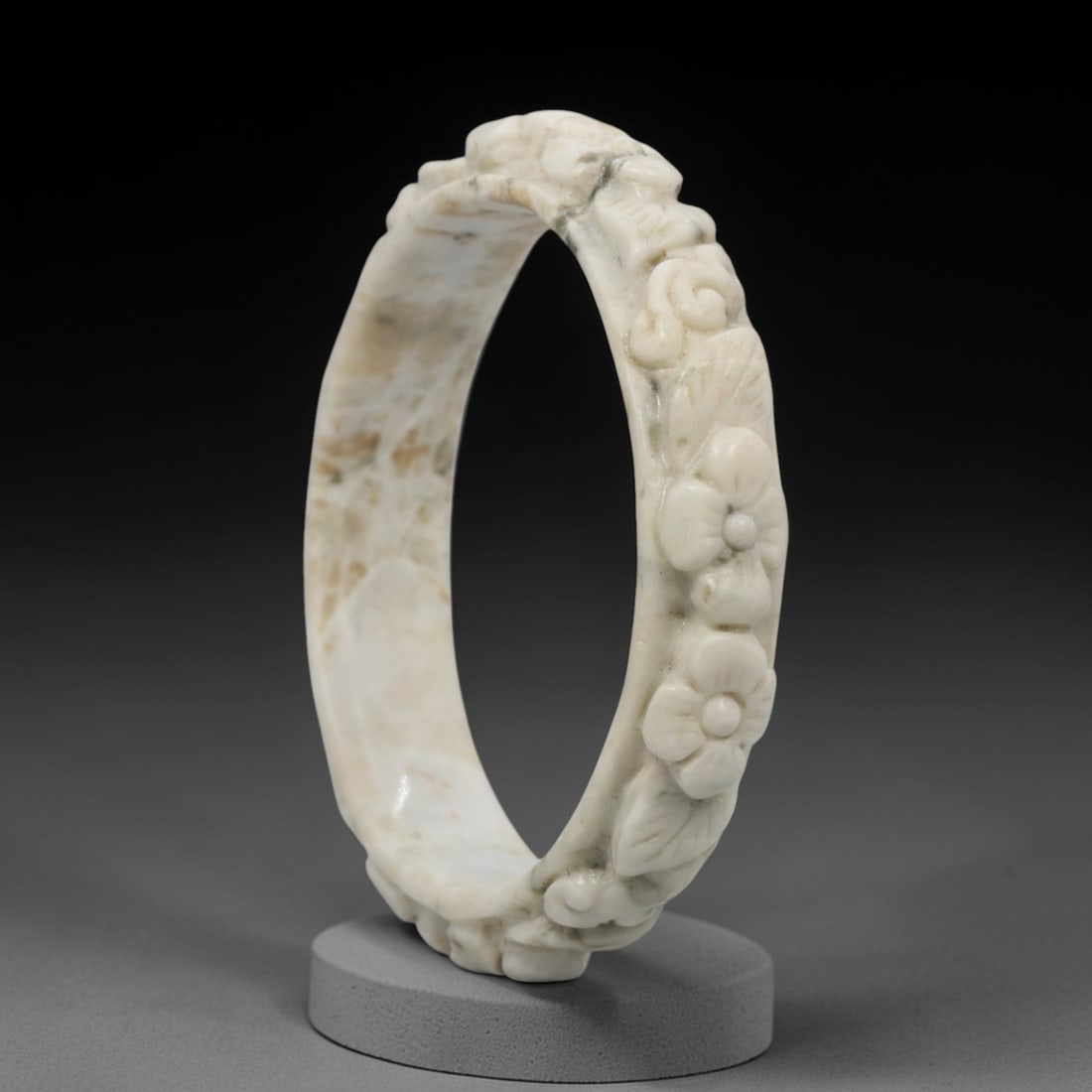 A Chinese Carved Jadeite Jade Bangle Bracelet, 20th Century, 75ZL: A fine jadeite bangle of flattened circular form, masterfully carved in relief with a continuous band of floral blossoms and scrolling foliate motifs. The stone exhibits a creamy white base, heavily a