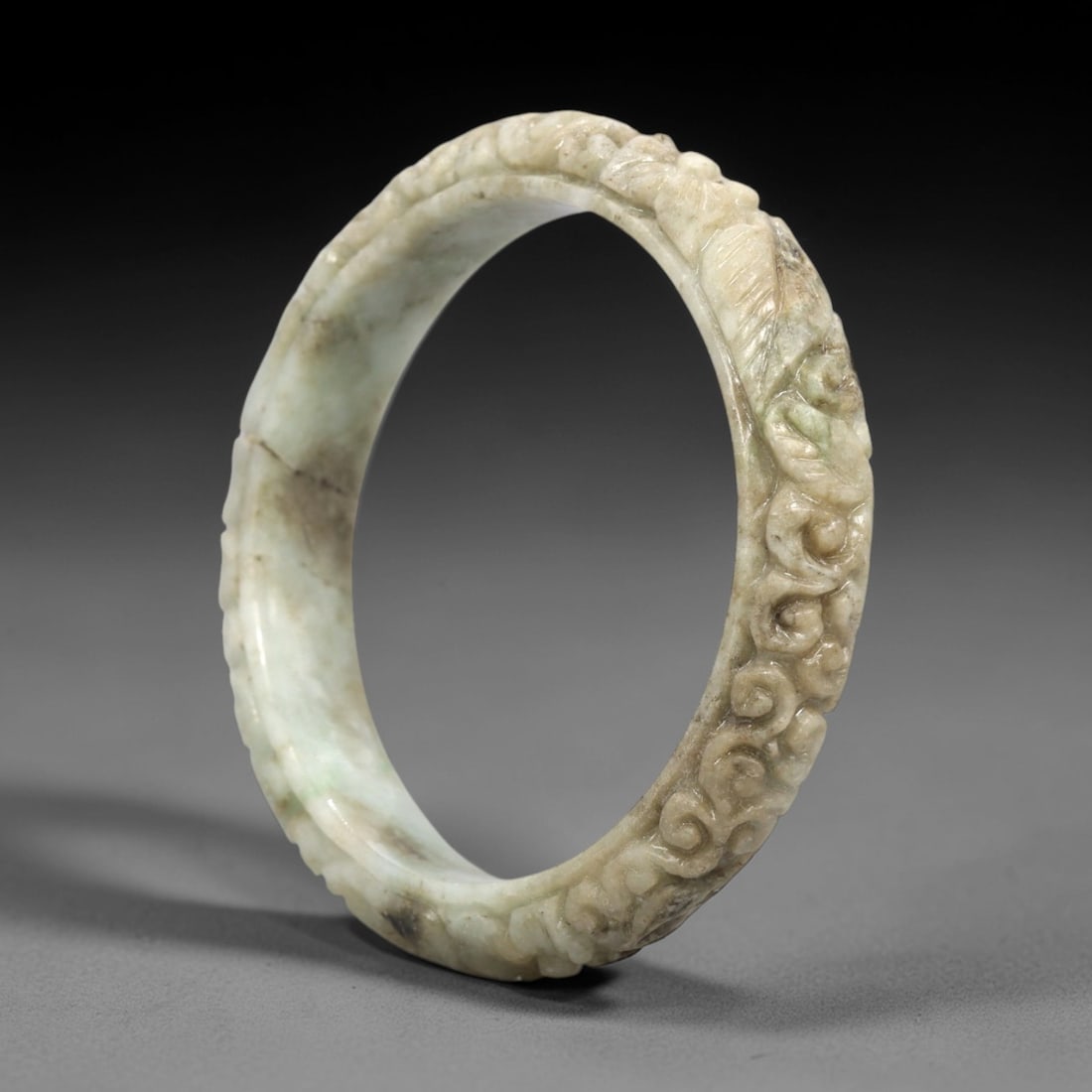 A Chinese Jadeite Jade Bangle Bracelet, 20th Century, 26FS: Carved from pale celadon jadeite jade with natural greyish and greenish tonal variation, the bangle is formed as a continuous circular band. The exterior surface is decorated with raised relief carvin