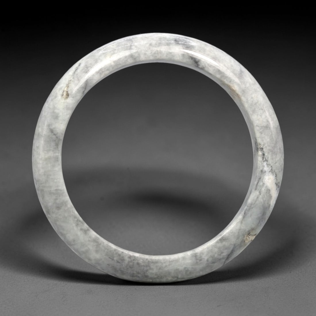 A Burmese Jadeite Jade Bangle, 20th Century, 15FS: A Burmese jadeite jade bangle, with a pale greyish-white matrix and scattered darker grey mineral inclusions. The bangle is of circular form, with a smooth, polished surface revealing the internal cry