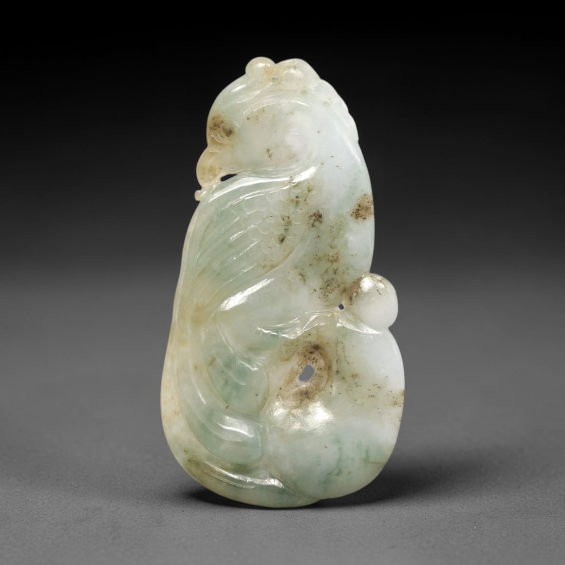A Chinese Jadeite Jade Phoenix Amulet Pendant, 20th Century, 92FR: Carved from translucent jadeite jade with a white base, pale green patches, and scattered brownish inclusions, the pendant is shaped as a stylized phoenix. The bird is rendered in relief, with a curve