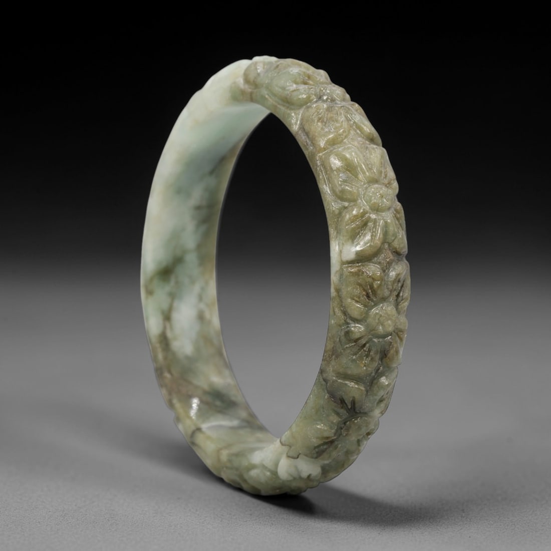 A Chinese Carved Jadeite Jade Bangle Bracelet, 20th Century, 72ZL: A fine jadeite bangle of flattened circular form, masterfully carved in relief with a continuous band of chrysanthemum blossoms. The stone exhibits a variegated palette, with a pale milky-green base t