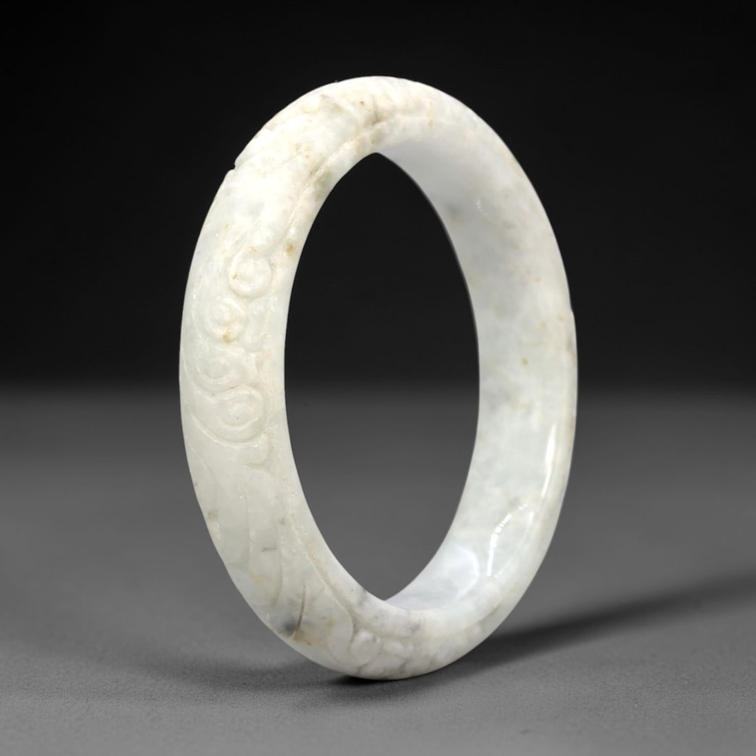 A Chinese Jadeite Jade Bangle Bracelet, 20th Century, 25FS: Carved from pale milky-white jadeite jade with subtle greyish and russet inclusions, the bangle is formed as a continuous circular band. The exterior surface is decorated with raised relief carving de