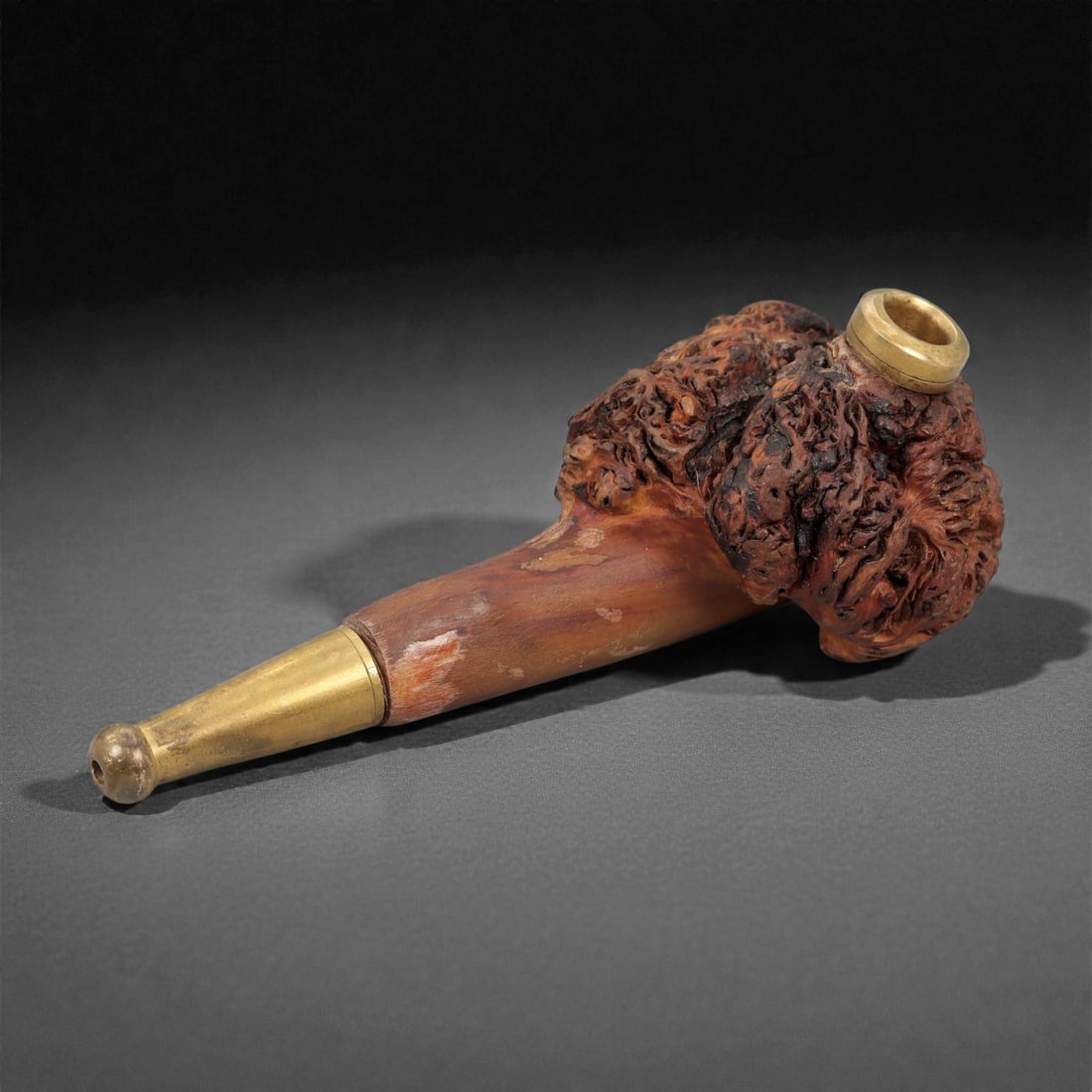 A Carved Wood Smoking Pipe, 20th Century, 23FH: Carved from hardwood, the pipe is composed of a compact bowl with a cylindrical interior and a tapered stem fitted with metal mounts. The bowl exterior is worked in high relief to follow the natural b