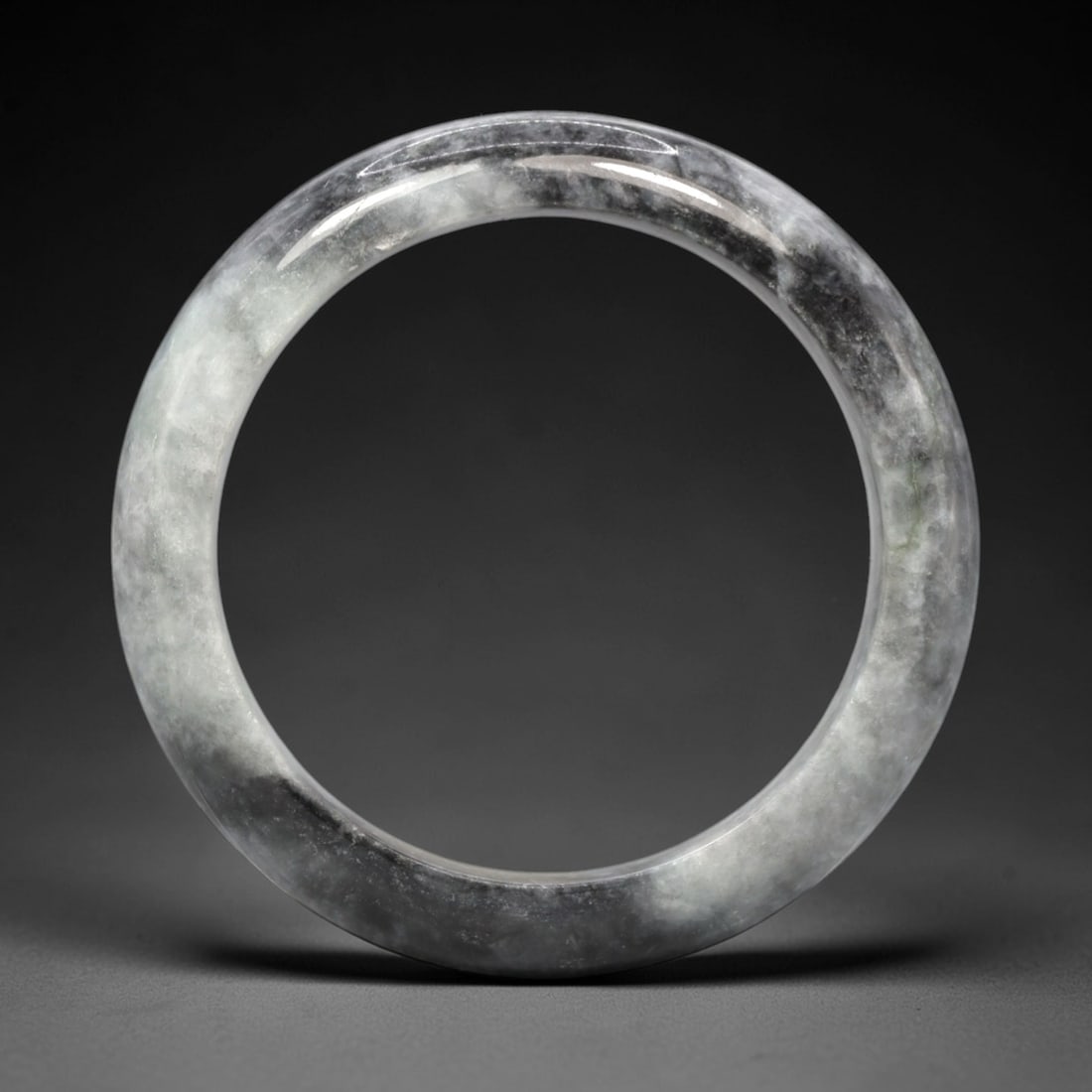 A Burmese Jadeite Jade Bangle, 20th Century, 1FS: A Burmese jadeite jade bangle, with a pale greyish-white matrix and prominent dark grey mineral inclusions. The bangle is of circular form, with a smooth, polished surface revealing the internal cryst
