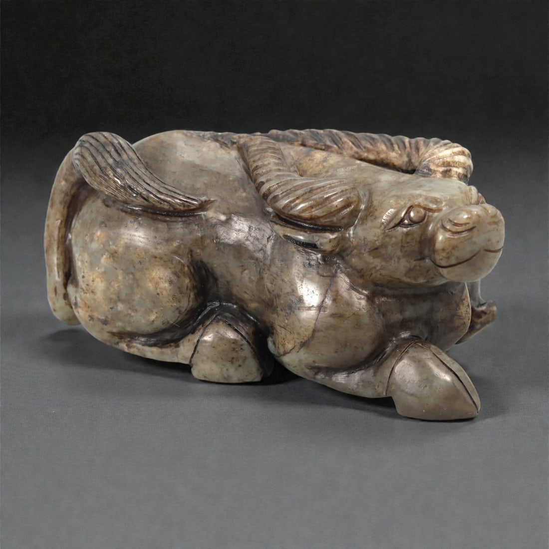A Chinese Jade Buffalo Sculpture, 20th Century, 34YA: Carved from mottled grey-green jade with areas of brown and dark mineral inclusions, the sculpture represents a recumbent water buffalo modeled fully in the round. The animal is shown resting with its