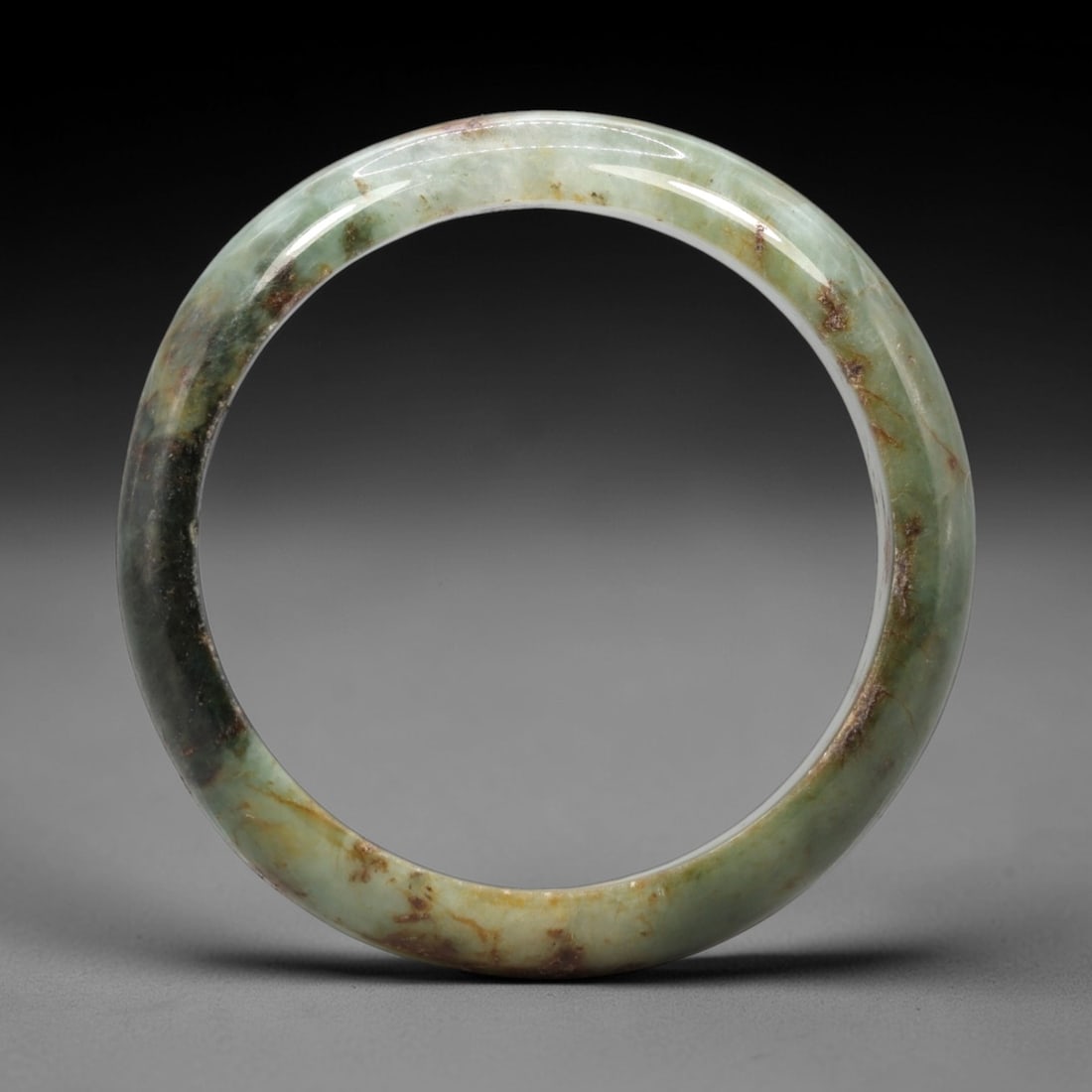 A Chinese Jadeite Jade Bangle Bracelet, 20th Century, 3ZL: Carved from jadeite jade with a pale green base and scattered russet-brown inclusions concentrated along the lower edge, the bangle is formed as a continuous circular band with a rounded cross-section