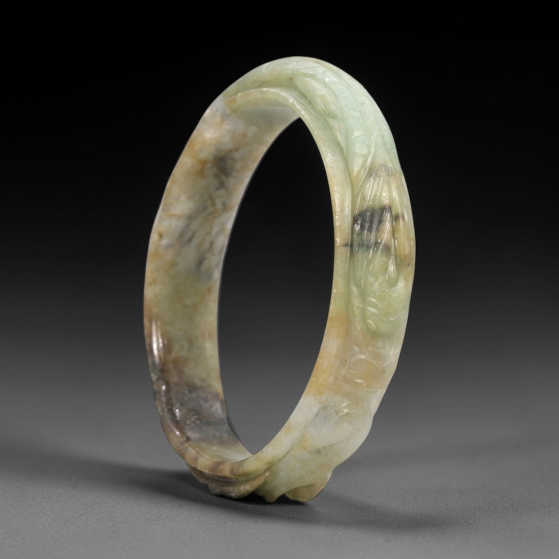 A Chinese Jadeite Jade Bangle Bracelet, 20th Century, 24FS: Carved from variegated jadeite jade with natural green, yellowish-brown, and dark inclusions, the bangle is formed as a continuous circular band. The exterior surface is decorated with raised relief c