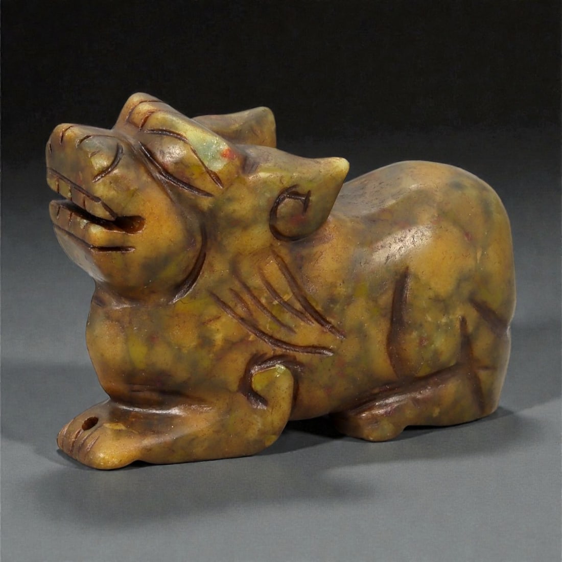 A Chinese Hardstone Pixiu Figure, Qing Dynasty, 19th Century, 17YB: Carved from mottled brown and yellow-toned hardstone, possibly soapstone, the figure represents pixiu, the mythical hybrid creature associated with wealth and protection. The animal is shown recumbent