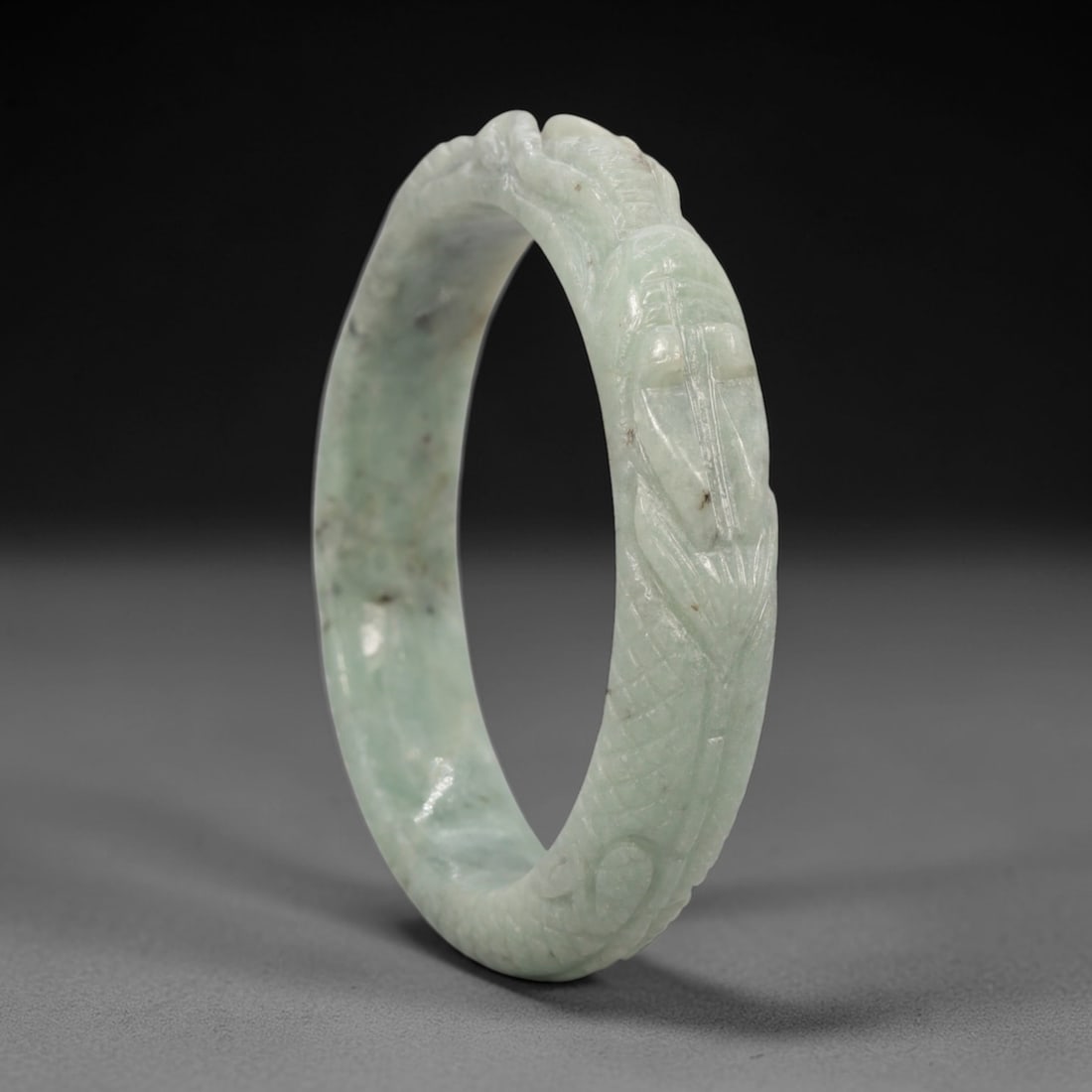 A Chinese Carved Jadeite Jade Bangle Bracelet, 20th Century, 74ZL: A fine jadeite bangle of circular form, masterfully carved in relief with a continuous band of a sinuous dragon coiling around its circumference. The stone exhibits a translucent pale green base, with