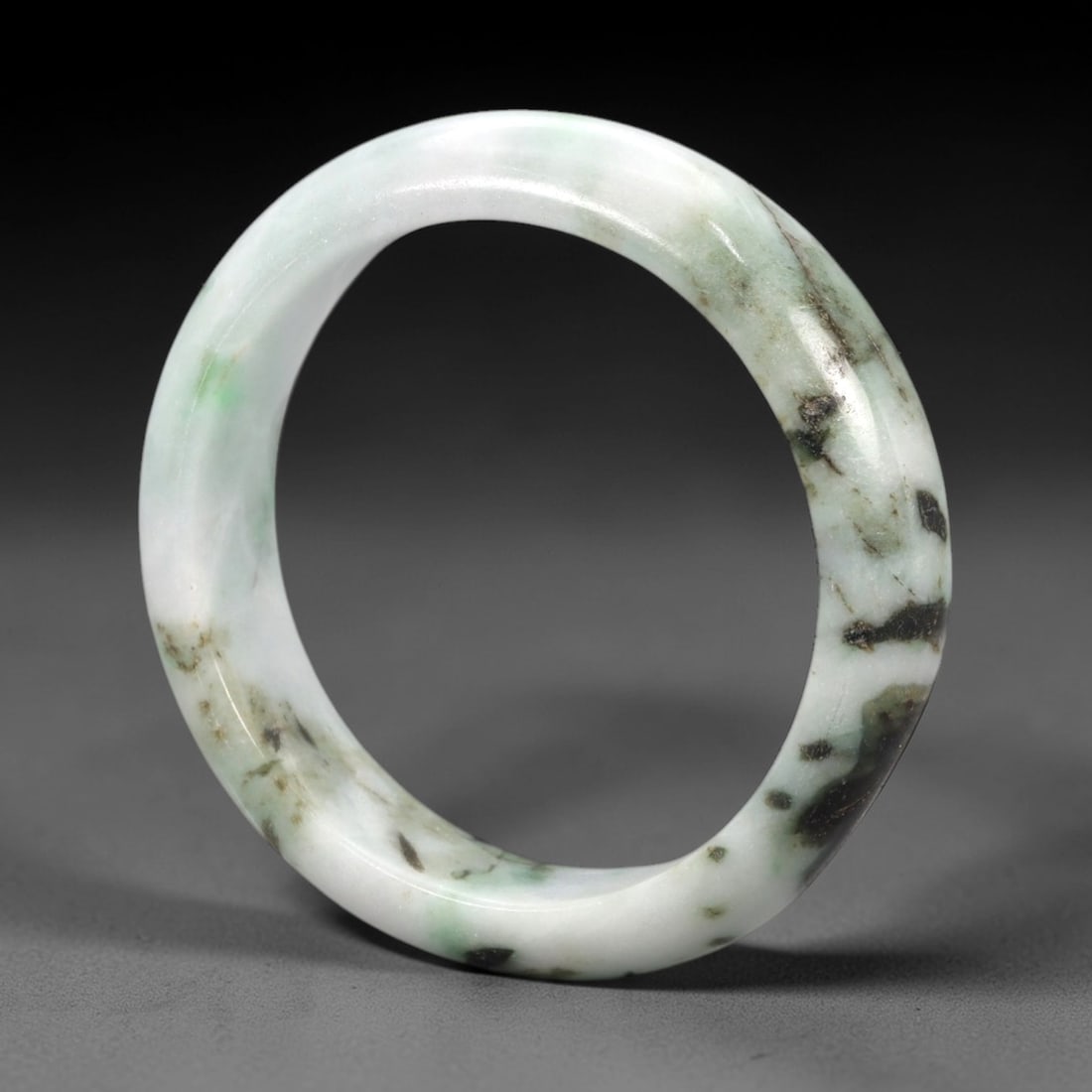A Chinese Jadeite Jade Bangle Bracelet, 20th Century, 88ZK: Carved from jadeite jade with a pale white base and scattered dark green and black inclusions, the bangle is formed as a continuous circular band with a rounded cross-section. The surface is smoothly