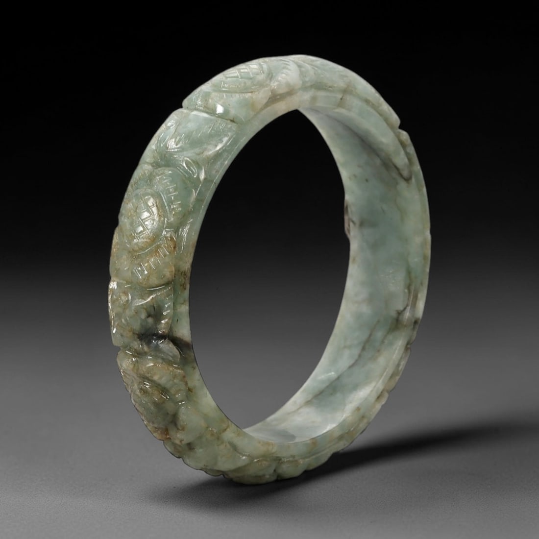 A Chinese Carved Jadeite Jade Bangle Bracelet, 20th Century, 36ZL: A fine jadeite bangle of flattened circular form, masterfully carved in relief with a continuous band of auspicious lingzhi and floral motifs. The stone exhibits a rich, translucent celadon-green base