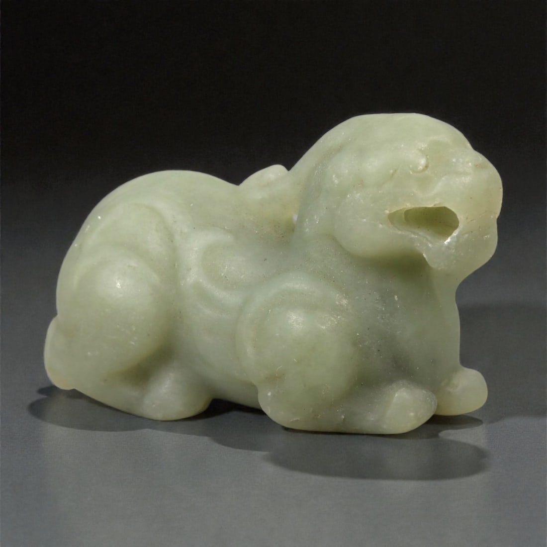 A Chinese Nephrite Jade Pixiu Figure, 20th Century, 84FA: Carved from pale celadon nephrite jade with soft translucency and scattered natural inclusions, the figure represents a pixiu shown recumbent with the limbs folded beneath the body. The head is slight