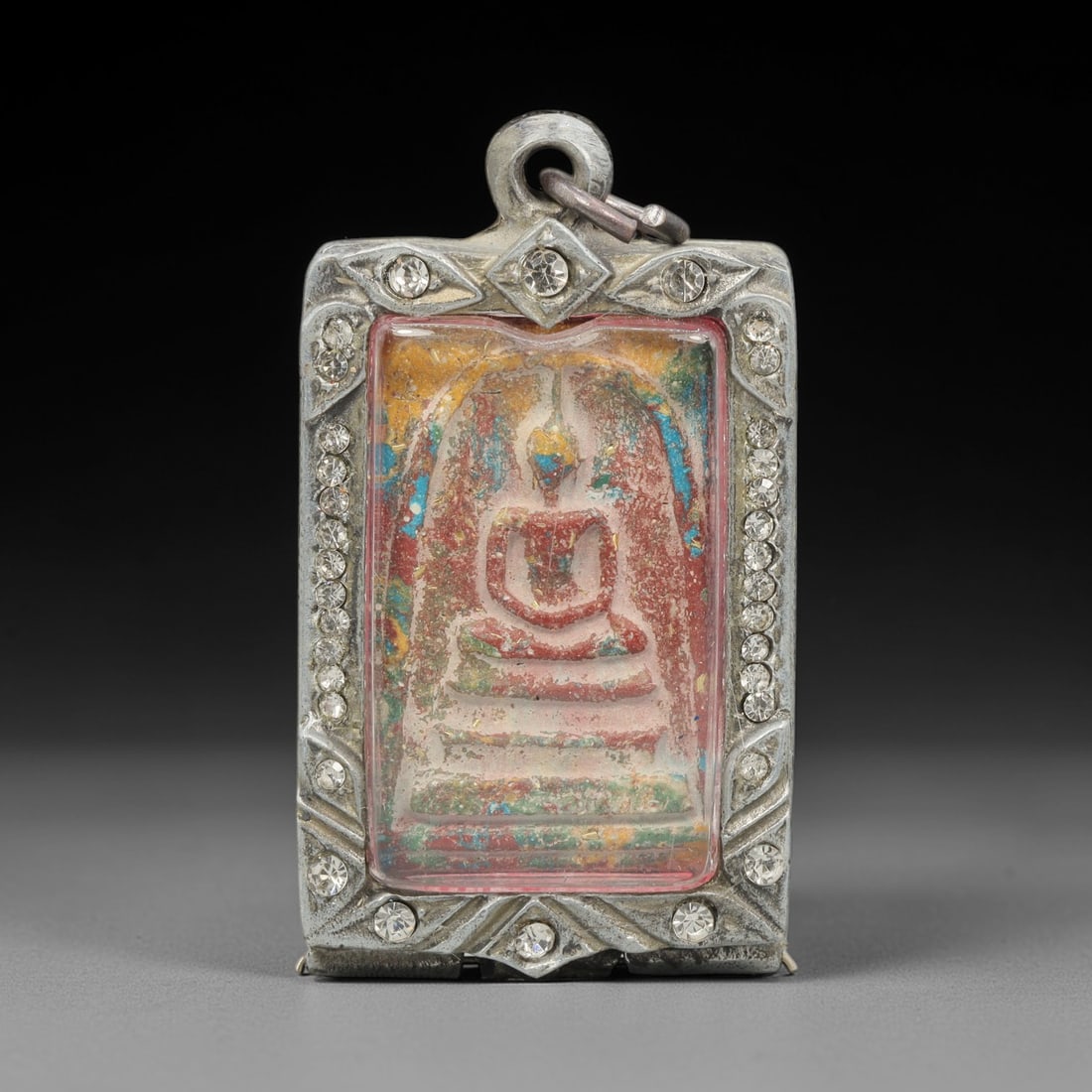 A Thai Terracotta Buddha Amulet in Metal Case, 20th Century, 84ZH: The amulet is formed from terracotta, with a matrix incorporating red, blue, yellow, and green pigmentation, creating a multi-colored surface. The obverse depicts a seated Buddha in bhumisparsha mudra