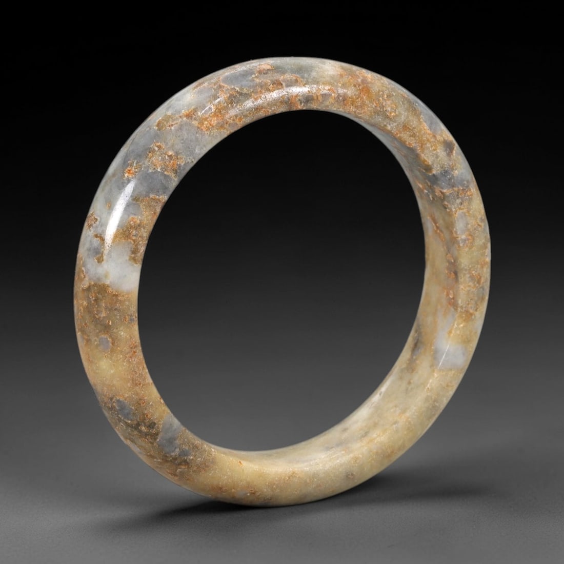 A Chinese Jadeite Jade Bangle Bracelet, 20th Century, 100ZK: Carved from jadeite jade with a pale yellowish-tan base and prominent russet and greyish-blue mineral inclusions concentrated along the upper edge, the bangle is formed as a continuous circular band w