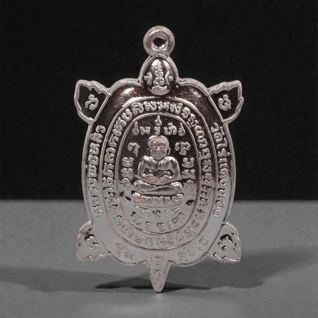 A Thai Silver-Toned Metal Turtle-Form Amulet, 21st Century, 73FP: Cast from metal with a silver-toned finish, the amulet is shaped as a turtle with an integral suspension loop at the apex. The obverse features a central seated Buddha figure in cross-legged meditatio