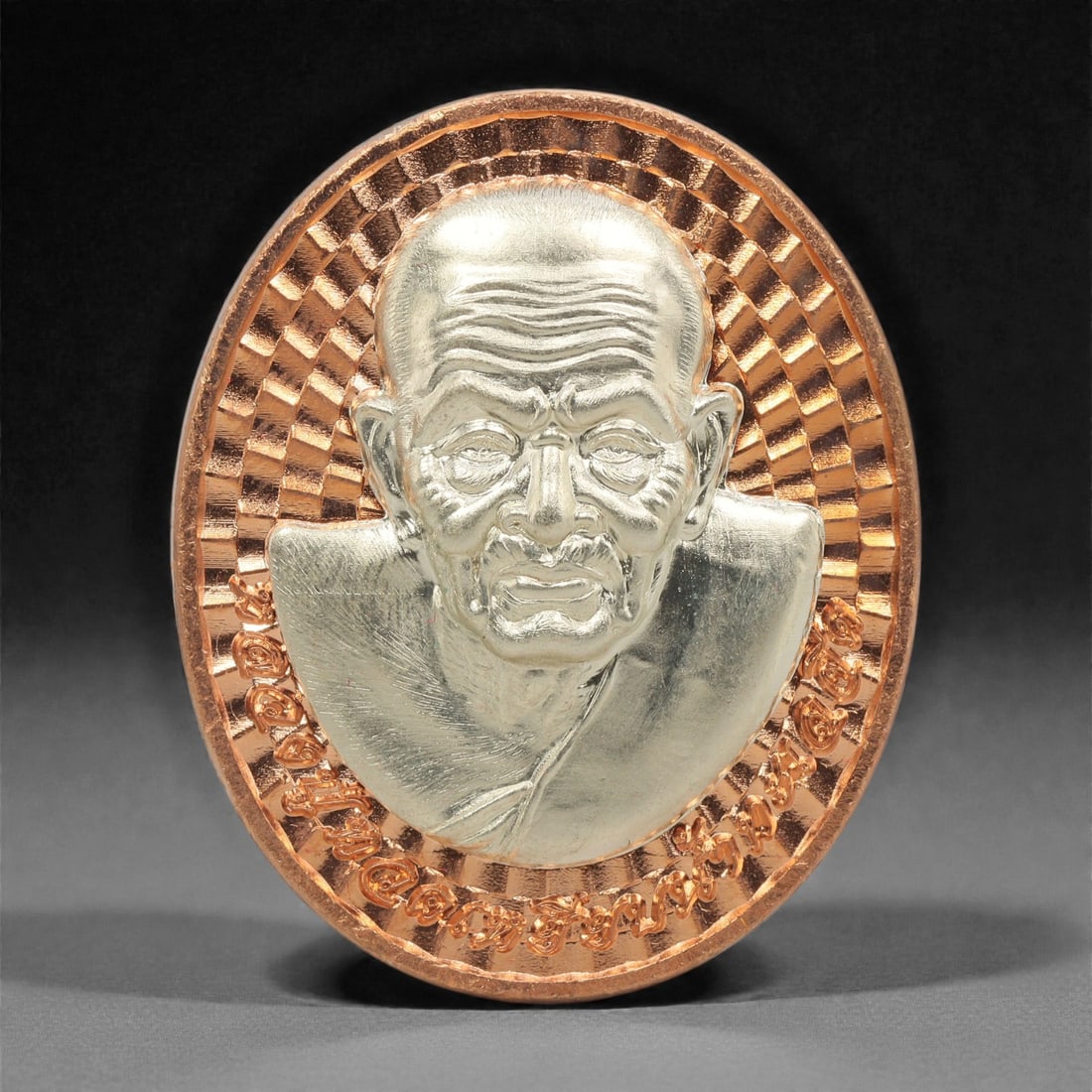 A Thai Bi-Metallic Monk Portrait Medallion, 21st Century, 89FP: Cast with a bi-metallic construction, the medallion is oval and double-sided with a raised copper-toned metal border. The obverse features a high-relief bust portrait of a Buddhist monk rendered in si