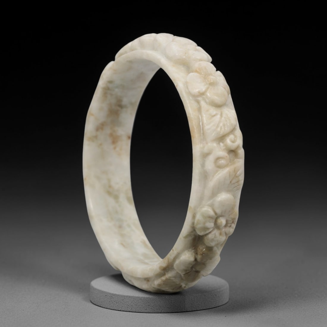 A Chinese Carved Jadeite Jade Bangle Bracelet, 20th Century, 29ZL: A fine jadeite bangle of flattened circular form, masterfully carved in relief with a continuous band of floral blossoms and scrolling foliate motifs. The stone exhibits a creamy white base, accented