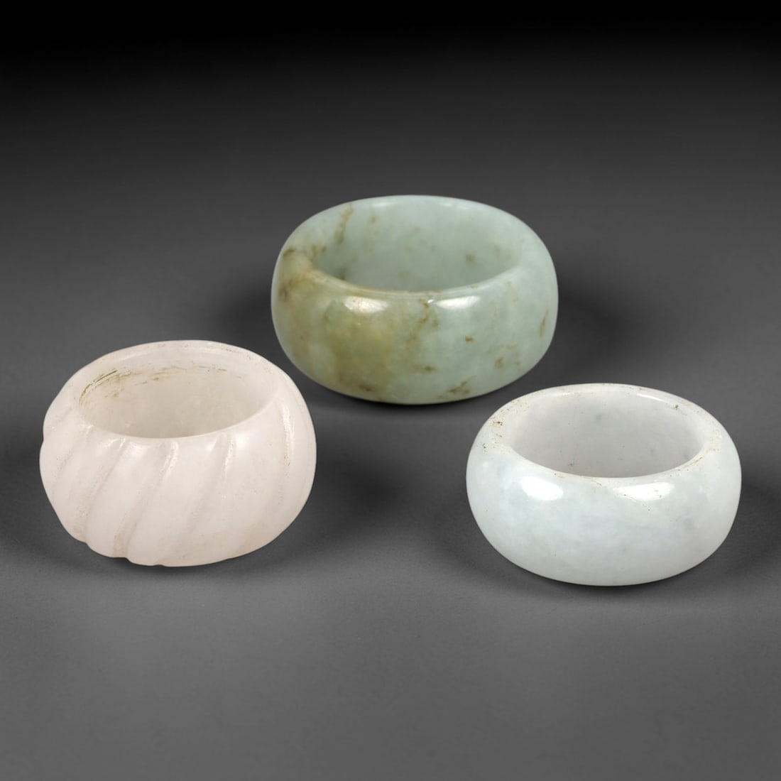 A Trio of Chinese Jadeite Rings - 20th Century, 26AL: This set of three Chinese jadeite rings features varied thick band forms crafted from natural jadeite in pale celadon, milky white, and white tonalities, with one ring exhibiting carved spiral fluting