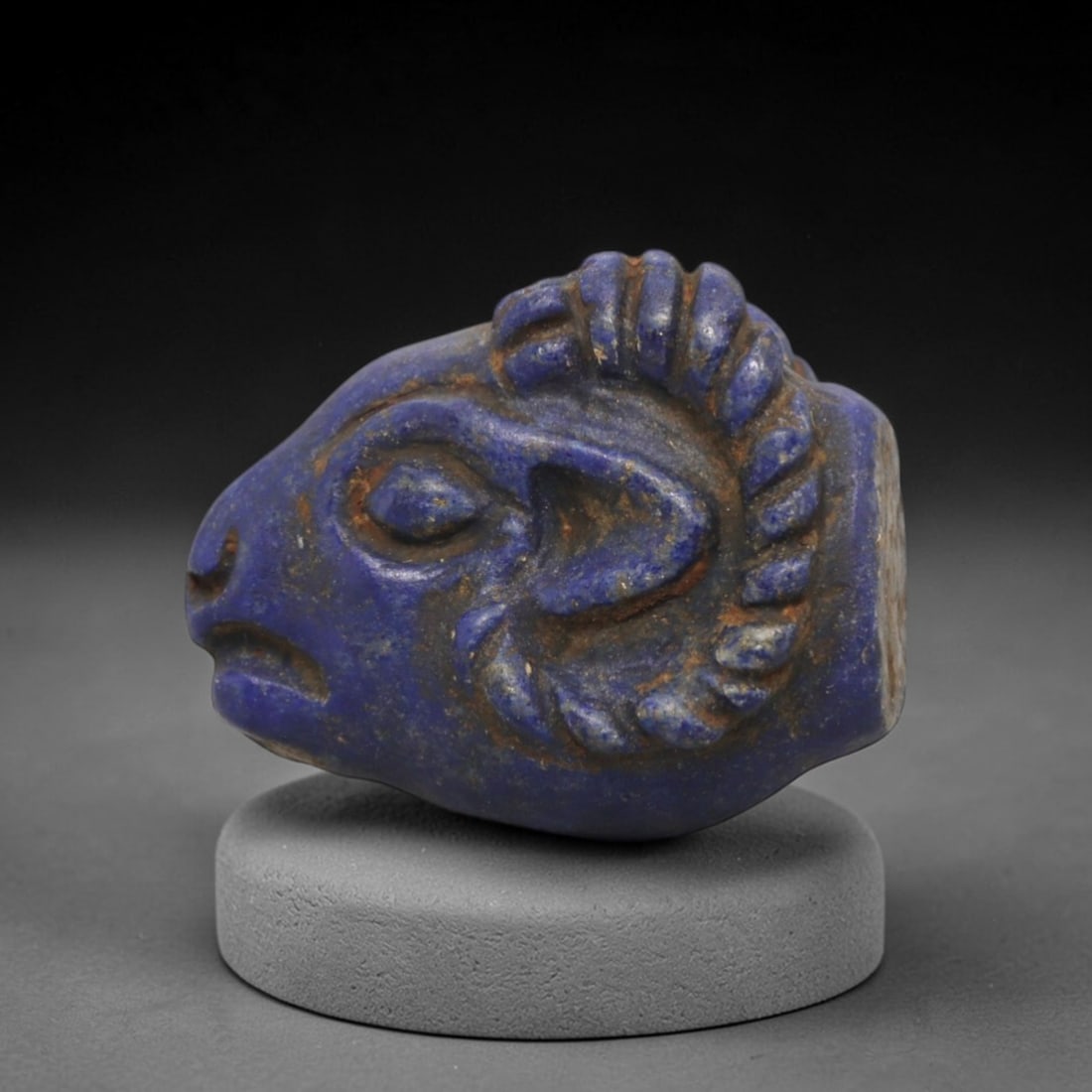 Ancient Lapis Lazuli Ram Head - Near Eastern Amulet Figurine, 99XV: Carved from lapis lazuli, this amulet depicts a stylized ram head with prominent, spiraled horns and detailed facial features. The eyes are deeply incised, the snout is defined, and the horns are rend